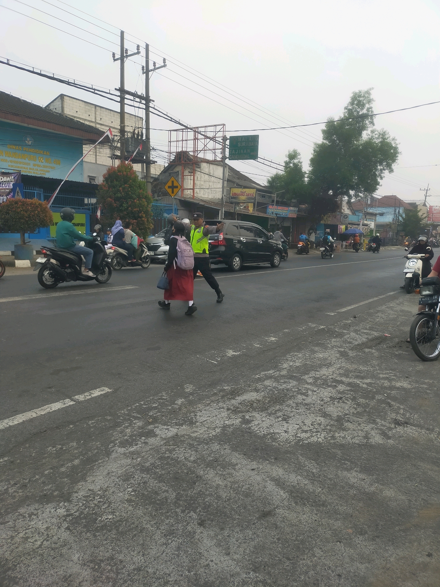The image depicts a street scene in Bululawang, Indonesia. A traffic controller, wearing a high-visibility yellow vest and dark pants, is directing traffic, holding up one hand. Beside him, a young female student in a red skirt and white shirt, carrying a light purple backpack, walks across the road. Behind them, a black car is stopped. On the left side of the road, a man in a green jacket and helmet rides a black motorcycle. Other motorcycles and pedestrians are visible in the background. The street is paved with asphalt and shows signs of wear. A building with blue signage, identified as "DINAS PENDIDIKAN" (Education Department), is visible on the left. Traffic signs and power lines are present overhead. The sky is overcast.