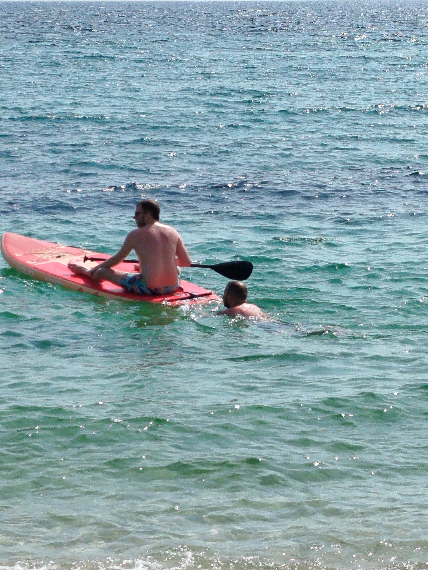 Two men are in the water off the coast of Sithonia Municipal Unit, Greece. One man is sitting on a red paddleboard, facing away from the camera and holding a paddle. He is wearing patterned swim trunks. The other man is partially submerged in the water, with his head and shoulders visible. The water is a clear, turquoise color and appears calm with small ripples. The sun is shining, creating bright reflections on the water's surface. The background shows a vast expanse of blue ocean meeting the horizon.