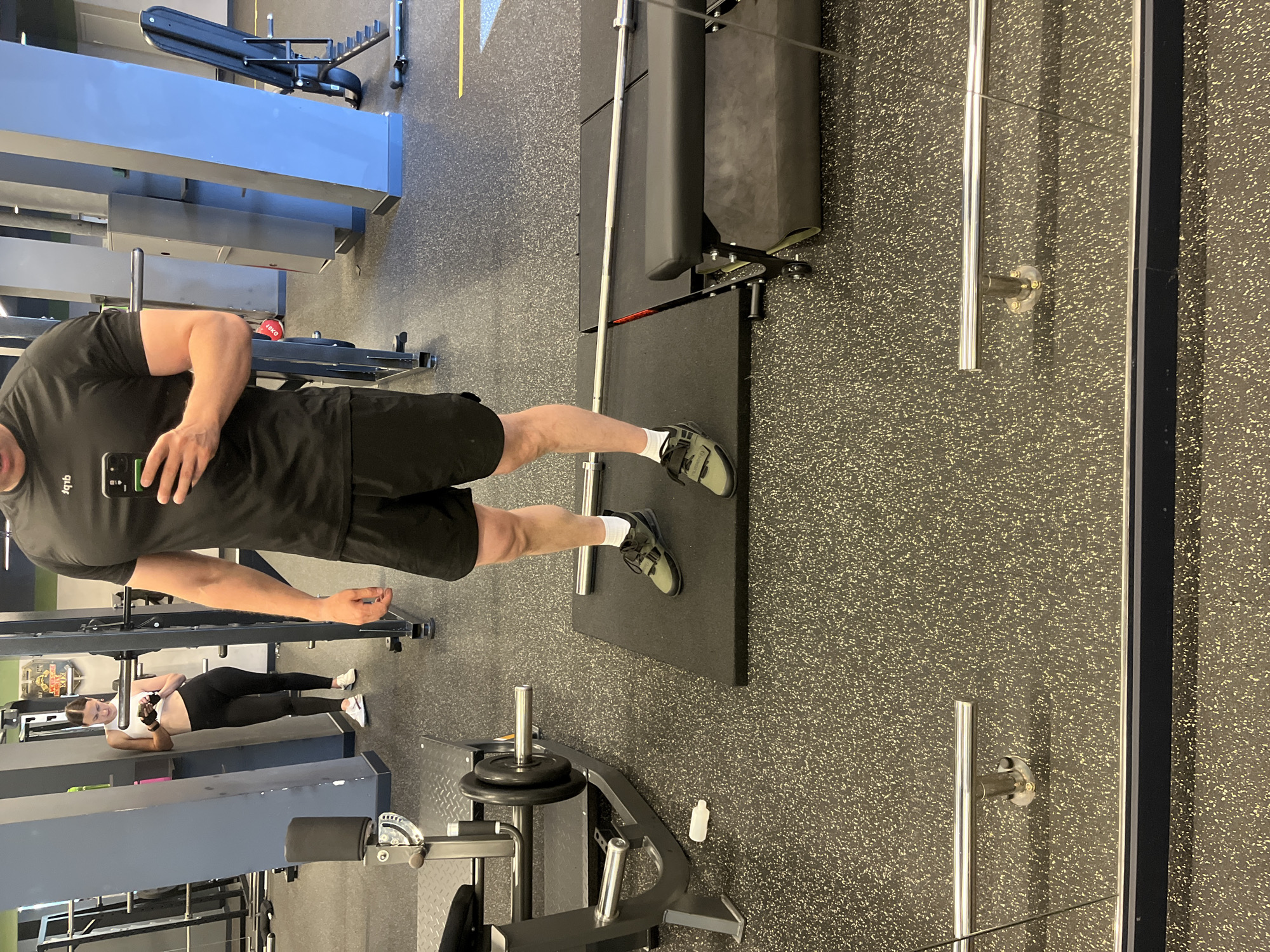 The image shows a man standing in a gym, holding his phone. He is wearing a black t-shirt and black shorts, with green athletic shoes and white socks. He is standing on a black mat. To his left, there is a weight bench with weights on it. In the background, a woman is standing and looking at her phone. She is wearing a white sports bra and black leggings, with white athletic shoes. The gym appears to be equipped with various weightlifting machines and equipment. The floor is covered with black rubber mats speckled with yellow. The overall environment suggests an indoor fitness facility. The presence of gym equipment and people engaged in fitness activities indicate a workout environment. The image does not provide any explicit visual cues to determine the specific location within Sochi, Russia.