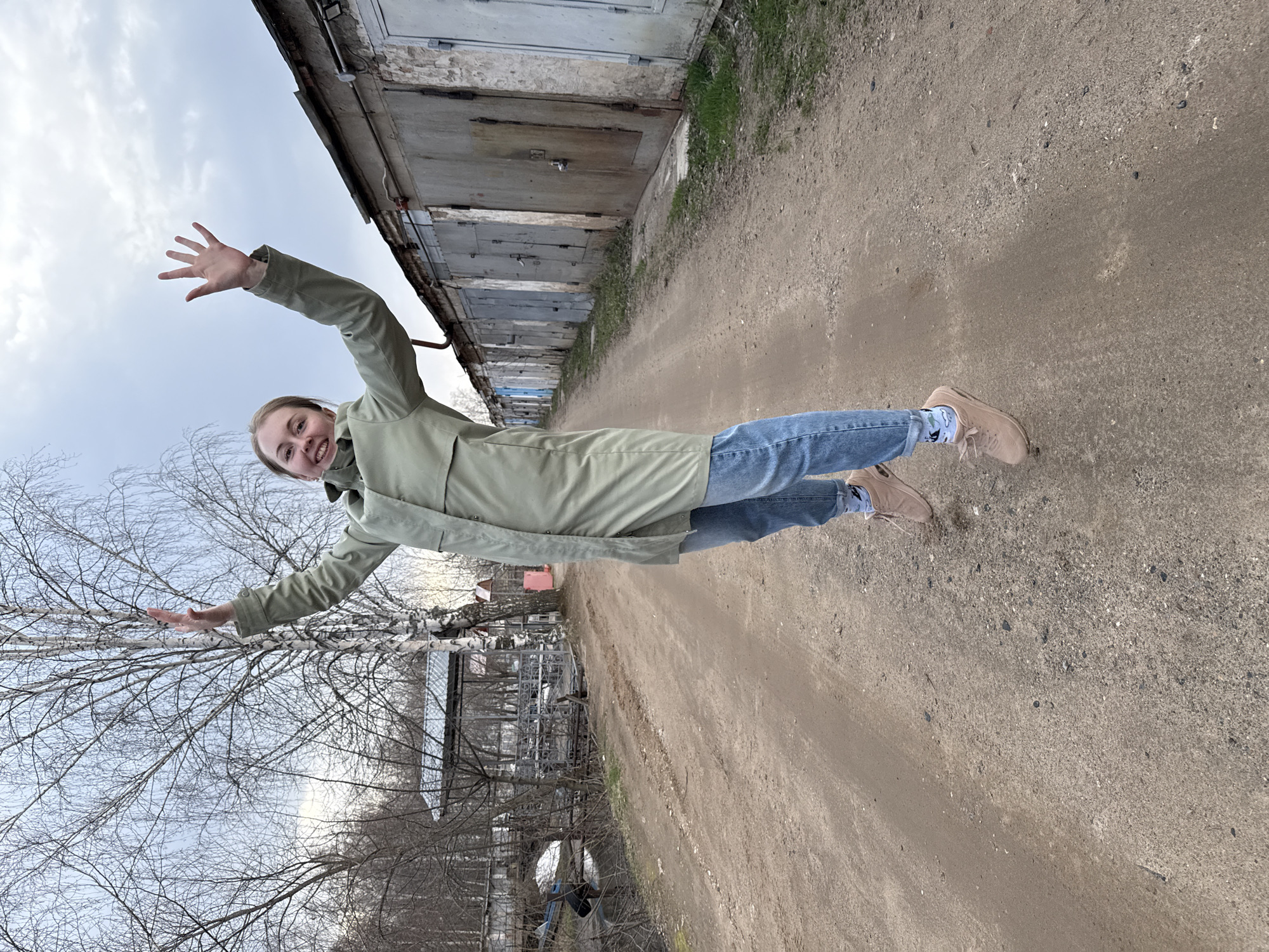 A young woman is captured mid-action in what appears to be a playful jump or pose, arms outstretched to her sides, against a backdrop of utilitarian structures and natural elements. She is smiling broadly, conveying joy and enthusiasm.
The woman wears a light green or khaki-colored long coat over blue jeans, paired with light brown lace-up shoes and patterned socks. Her light-colored hair is pulled back.
The setting features a dirt or gravel path underfoot, extending into the distance. To the right, a long row of weathered, grey garage or storage doors with some visible brickwork above lines the path. Patches of green grass grow at the base of these structures. To the left, several bare-branched trees, possibly birches, stand against a cloudy sky, indicating late winter or early spring. Hints of distant structures are visible through the trees.
The time of day appears to be daylight, under an overcast sky, contributing to a soft, even light. There is no visible text in the image. The overall scene suggests an outdoor, perhaps urban or suburban, backlot or pathway.