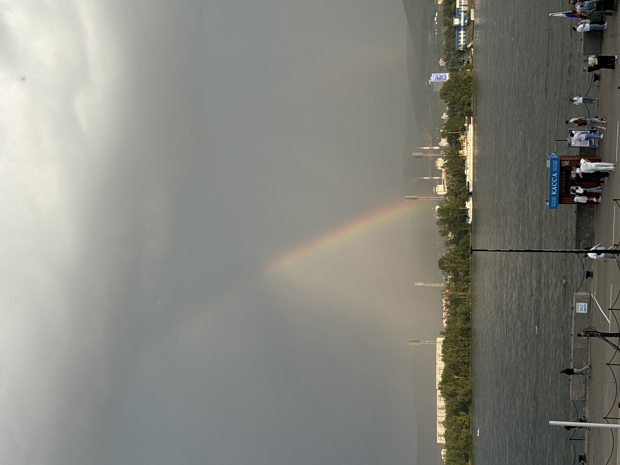 The image depicts a waterfront scene in Krasnoyarsk, Russia, likely on the Yenisei River. In the sky, a prominent rainbow arcs across a dramatic, cloud-filled backdrop, hinting at recent or ongoing rainfall. In the distance, across the wide river, a cityscape is visible, characterized by industrial structures with tall chimneys, a large residential or office building, and lush green trees. A few boats are present on the water, and a long, white vessel is docked.
In the foreground, a paved walkway lines the riverbank, with a low barrier and rope cordoning off the edge. Several people are scattered along this walkway, some standing, others walking. To the right, a small blue and white kiosk with the word "КАССА" (Cashier) printed on it is visible, suggesting it might be a ticket or information booth. A few individuals are gathered around this kiosk. A Russian flag is also visible on a flagpole near the kiosk. The overall atmosphere is somewhat somber due to the cloudy sky, but the rainbow provides a striking visual contrast.