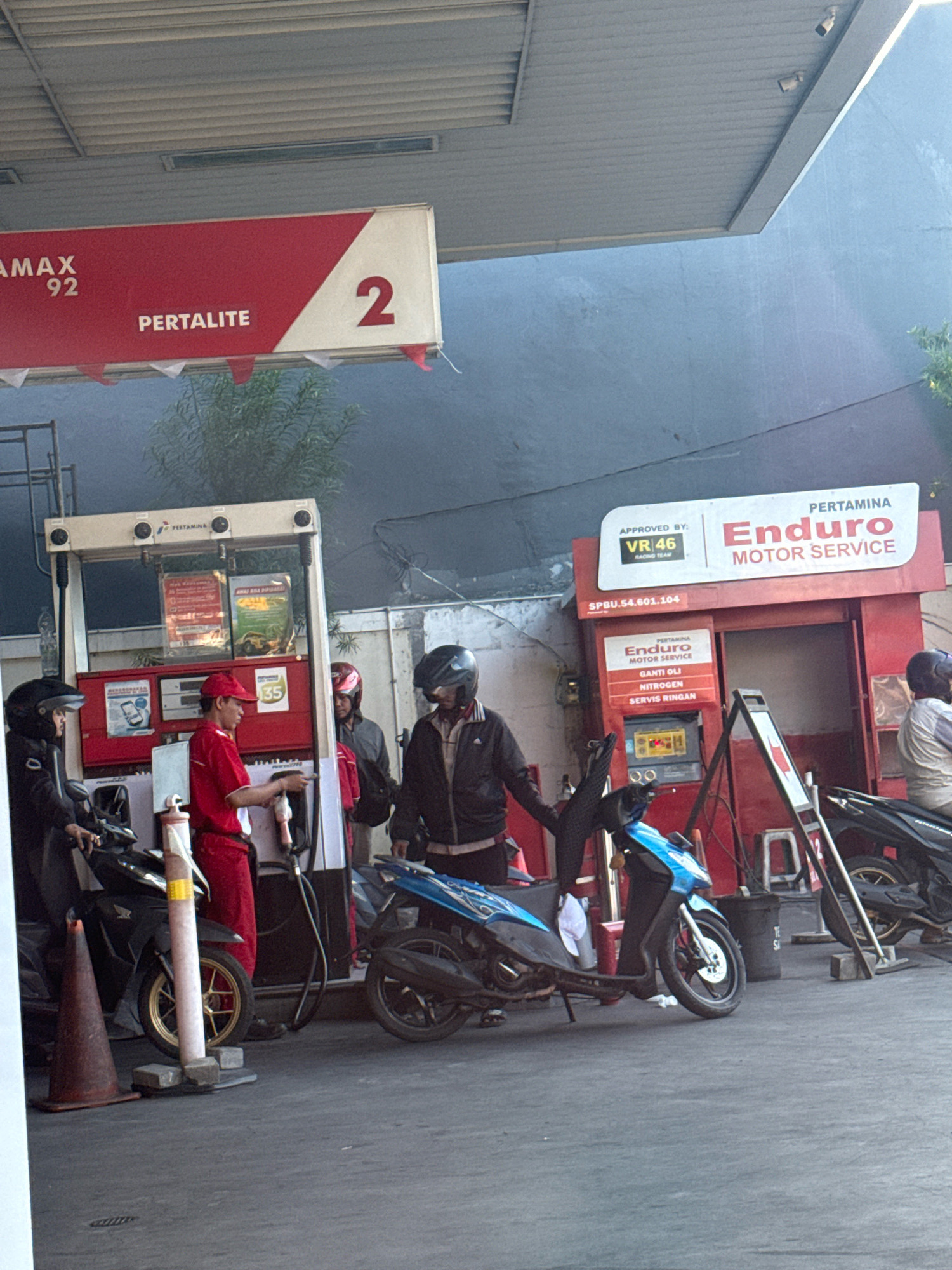 This is an eye-level, medium shot photograph taken at a Pertamina gas station in Surabaya, Indonesia. The scene is set during the day, under an awning of the gas station. In the foreground, there are two motorcycles, one black and one blue, parked on an asphalt surface. There are also three individuals present. One individual, wearing a red uniform and a red cap, is interacting with a gas pump. Another individual, wearing a black jacket and helmet, is standing next to the black motorcycle. A third individual, also wearing a helmet, is seated on the blue motorcycle. In the background, there is a red building with signs that read "PERTAMINA" and "Enduro MOTOR SERVICE." The overall scene depicts a typical transaction at a gas station.