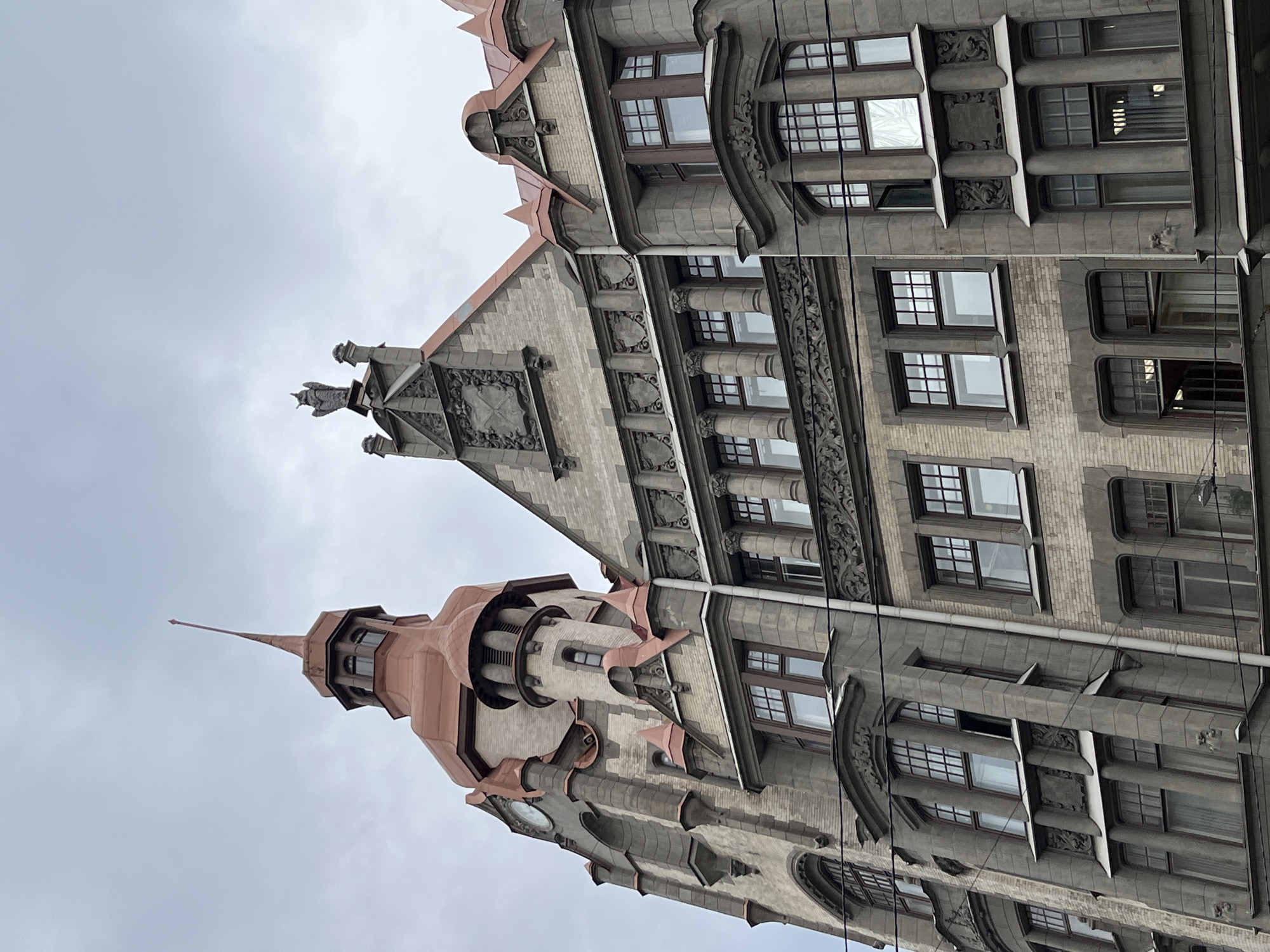 The image depicts the architectural details of two interconnected buildings in Saint Petersburg, Russia. The predominant architectural style features elements of Art Nouveau and Gothic Revival, with ornate detailing, including decorative stonework, turrets, and gables. The building on the left features a tall, slender turret with a copper-colored spire. The building on the right showcases rows of windows with decorative surrounds. The sky is overcast, contributing to the somber atmosphere. There are no people visible in the image.