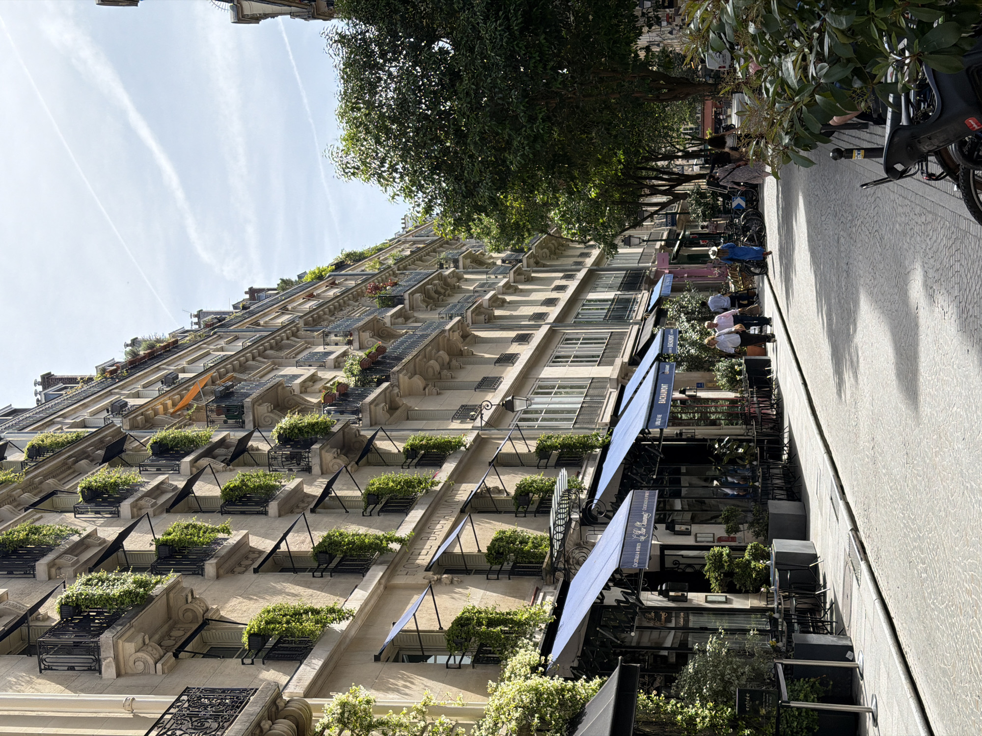 The image depicts a street scene in Paris, France. A tall building with many balconies and window boxes filled with greenery dominates the left side of the frame. Awnings are extended from the building, casting shadows on the sidewalk. On the right is a paved walkway with people strolling and bicycles parked. A row of trees lines the street, providing shade. The sky is visible at the top, with some light clouds. The overall impression is of a typical Parisian street, combining residential architecture with urban life.