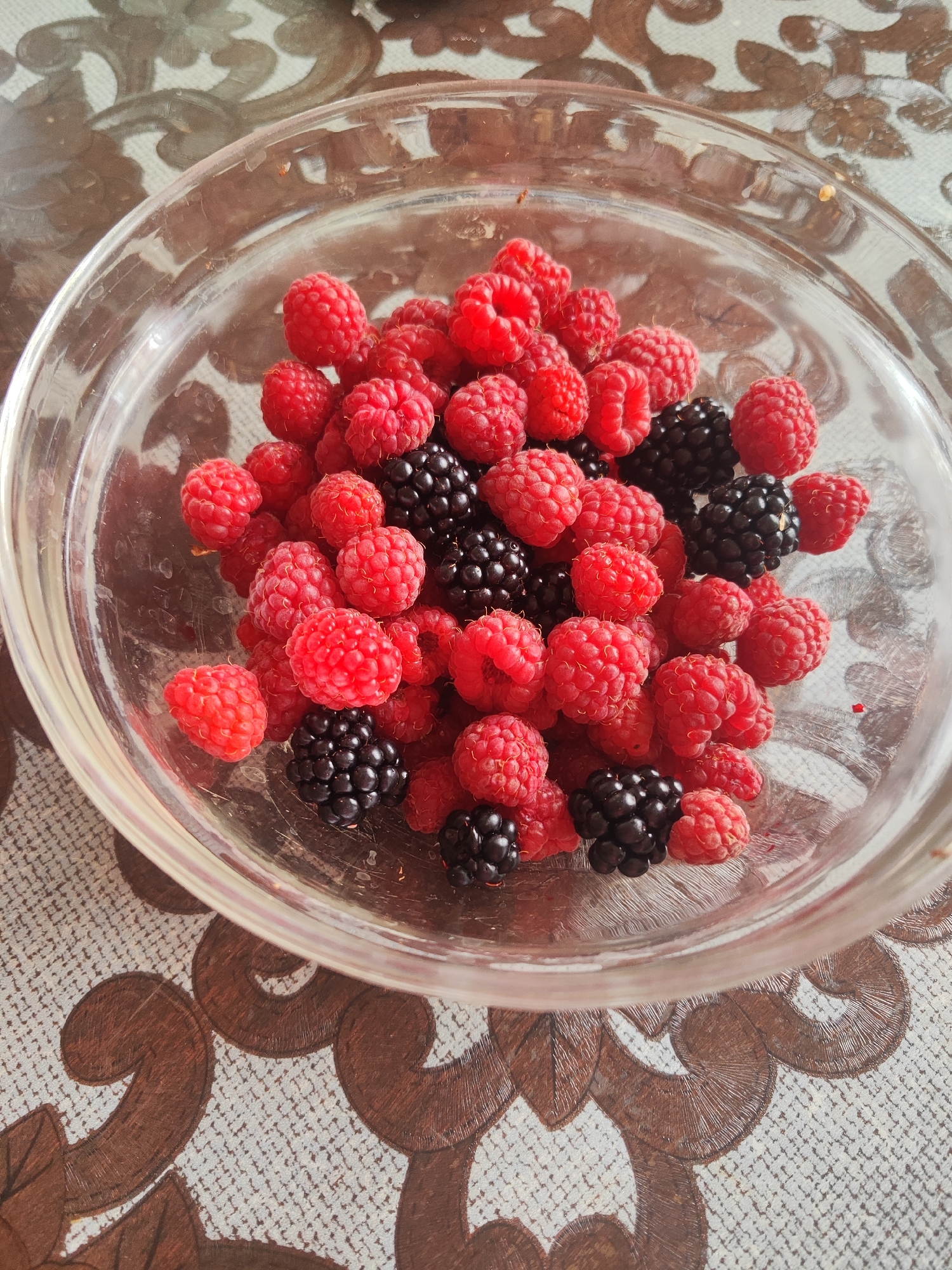 A close-up, overhead shot showcases a clear glass bowl filled with a mix of ripe raspberries and blackberries, resting on a textured placemat with a brown floral pattern. The berries are fresh and vibrant, with raspberries being predominantly red and some blackberries appearing black and shiny. The lighting suggests an indoor setting, possibly on a table. The image does not contain any identifiable people or specific landmarks that would indicate its location in Gvardeysk, Russia.