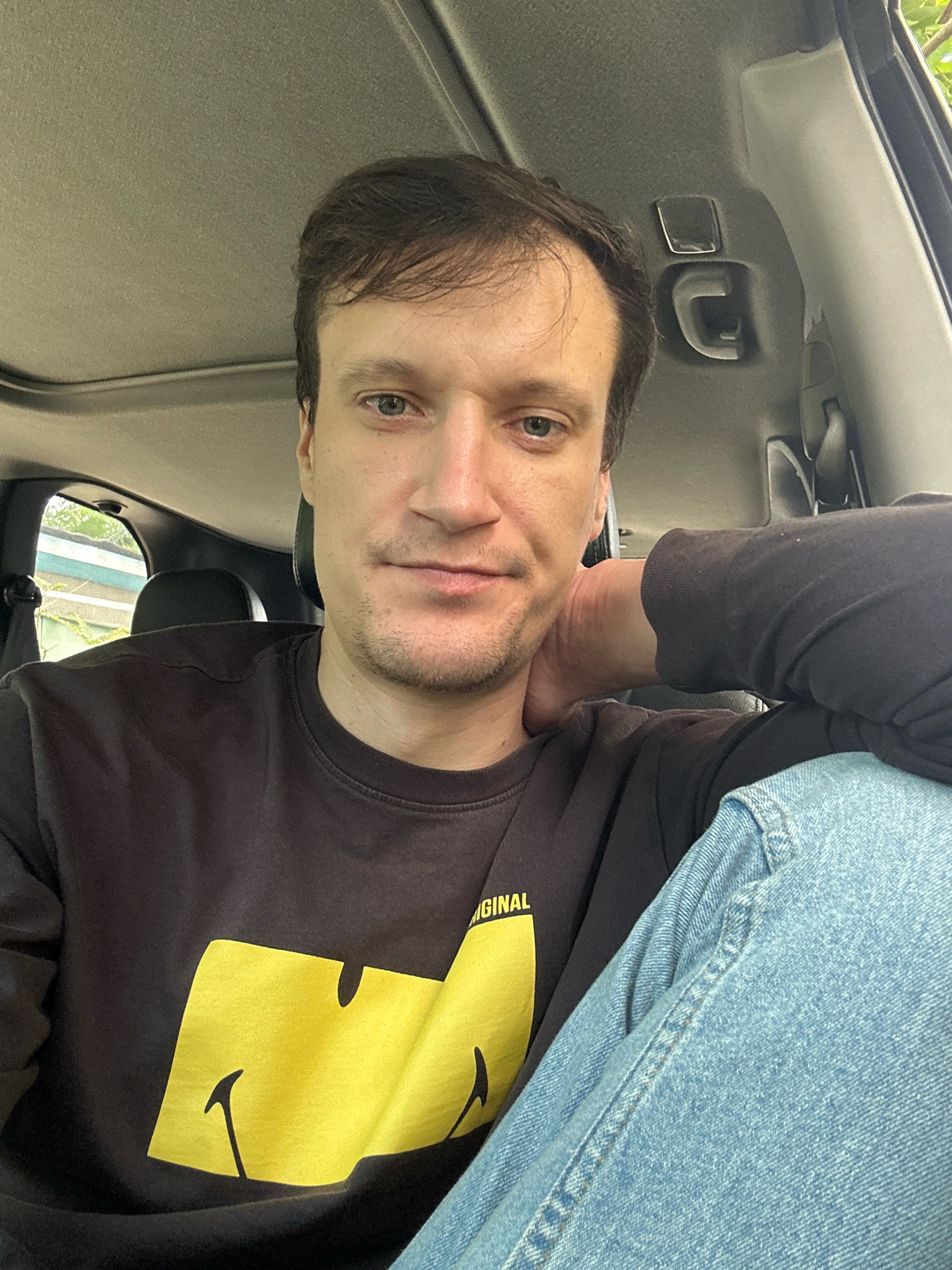 A young adult male with brown hair and blue eyes is seated in the driver's seat of a vehicle, likely a car, with his hand resting on the back of his neck. He is wearing a dark brown long-sleeved shirt with a large yellow smiley face graphic and the word "ORIGINAL" printed on it, along with light blue denim jeans. The interior of the vehicle is visible, including the ceiling with a sunroof, a grab handle, and the headrest of a passenger seat. The background outside the car window suggests an outdoor environment, possibly with some greenery, but the specific location in Makiv, Ukraine, cannot be determined from the image alone. The lighting indicates it is daytime.
