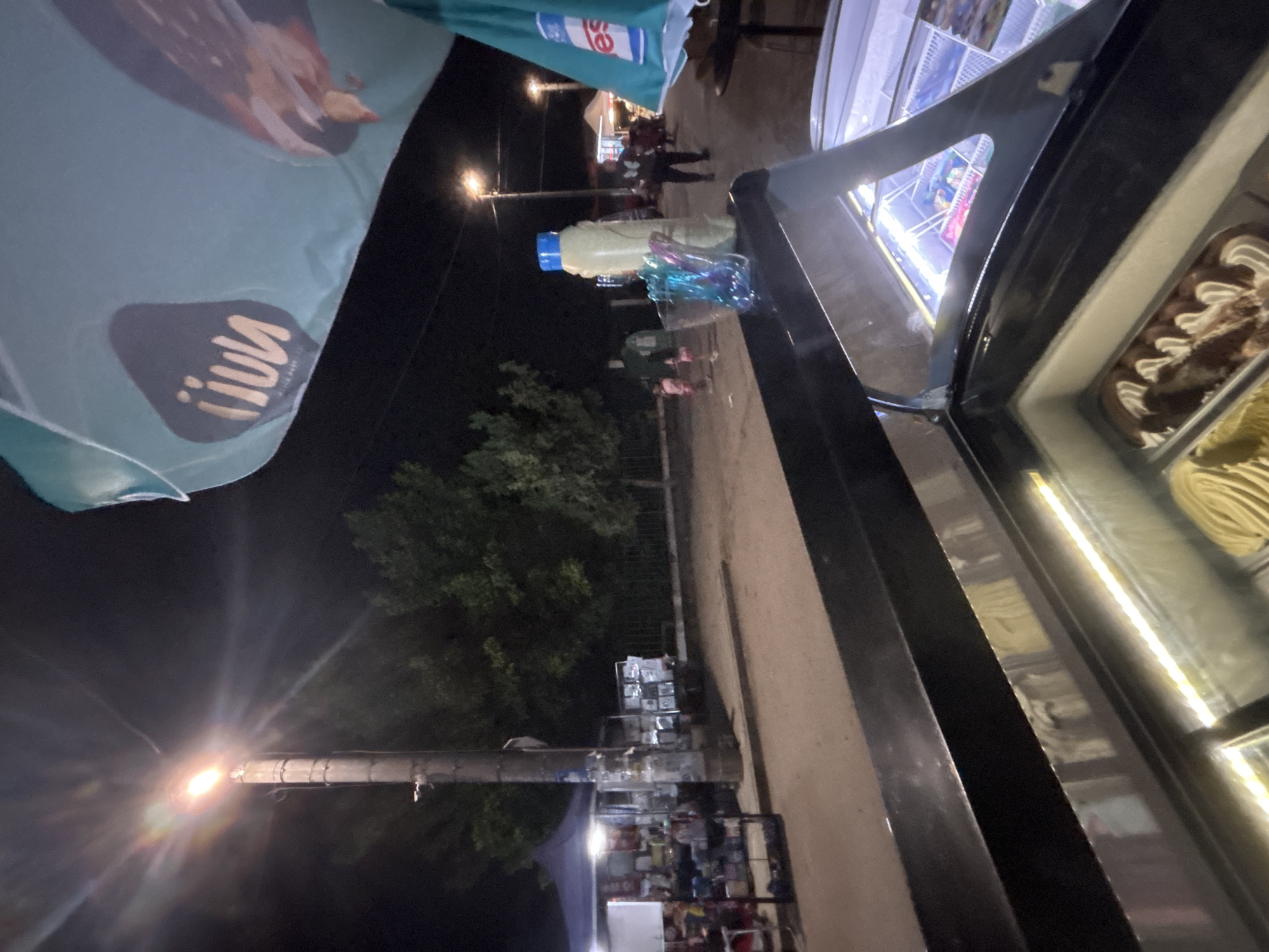 The image depicts an outdoor, nighttime scene in the city of Kostievo, Bulgaria, likely a street market or fair. In the immediate foreground, a black-framed ice cream display freezer is visible, with its glass lid open. Inside, multiple containers of ice cream are present, including one with chocolate and white cream swirls, and another with a yellow, solid appearance. The interior of the freezer is illuminated by bright linear LED lights.
Resting on the black frame of the ice cream display, to the right, are a tall, opaque, light-yellow bottle with a blue screw cap, and a clear glass or plastic holder containing multiple colorful plastic spoons or stirrers.
Above and to the right, a large portion of a light-blue or teal market umbrella is visible, featuring white scalloped edges. On its surface, a dark logo with the inverted text "iiuv" (likely "nuun" when viewed correctly) is present. Another partially visible logo, showing "es", is on the lower right section of the umbrella.
The midground features an unpaved or dirt road/path illuminated by various light sources. A prominent utility pole stands centrally on the left, topped with a bright, glowing street lamp emitting significant light flares. To the left of the pole, a vendor stall is set up under a dark blue tent, displaying various items for sale. To the right of the pole, another smaller display or stall is present.
Further down the path, several individuals are visible. Two smaller figures, likely children, one in a pink dress, are walking away from the camera, near a taller, dark-clothed adult figure. In the background, additional street lamps provide further illumination, and more people are discernible along the path. Dense, dark green trees and foliage line the background, indicating a natural or planted area. The sky is dark, consistent with the nighttime setting.