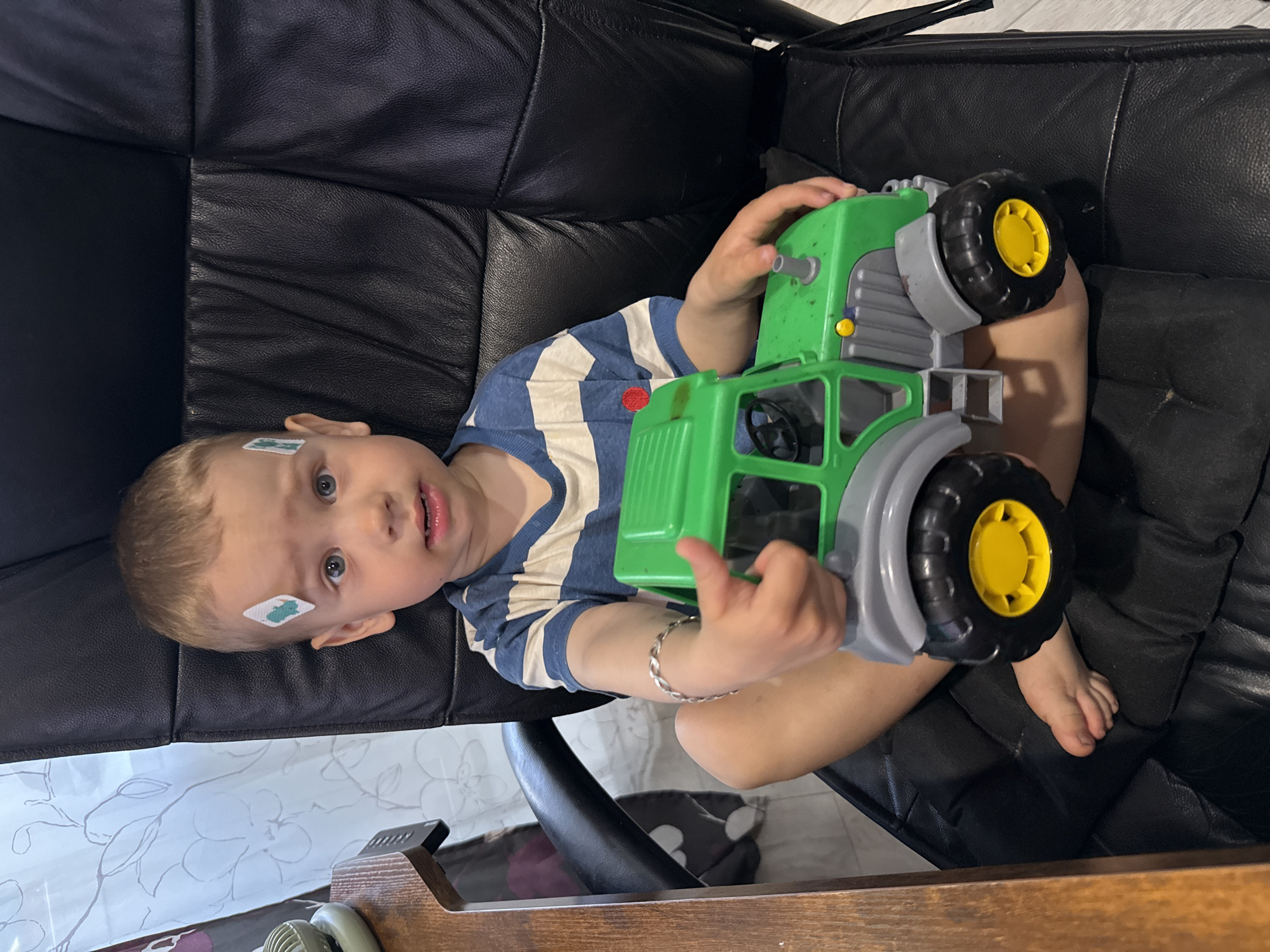 A young child lies on a black leather couch. The child has light hair and a bandage on their forehead. They wear a striped shirt and a silver bracelet. The child holds a green and gray toy tractor. The location is suggested to be Rusanovo, Russia.
