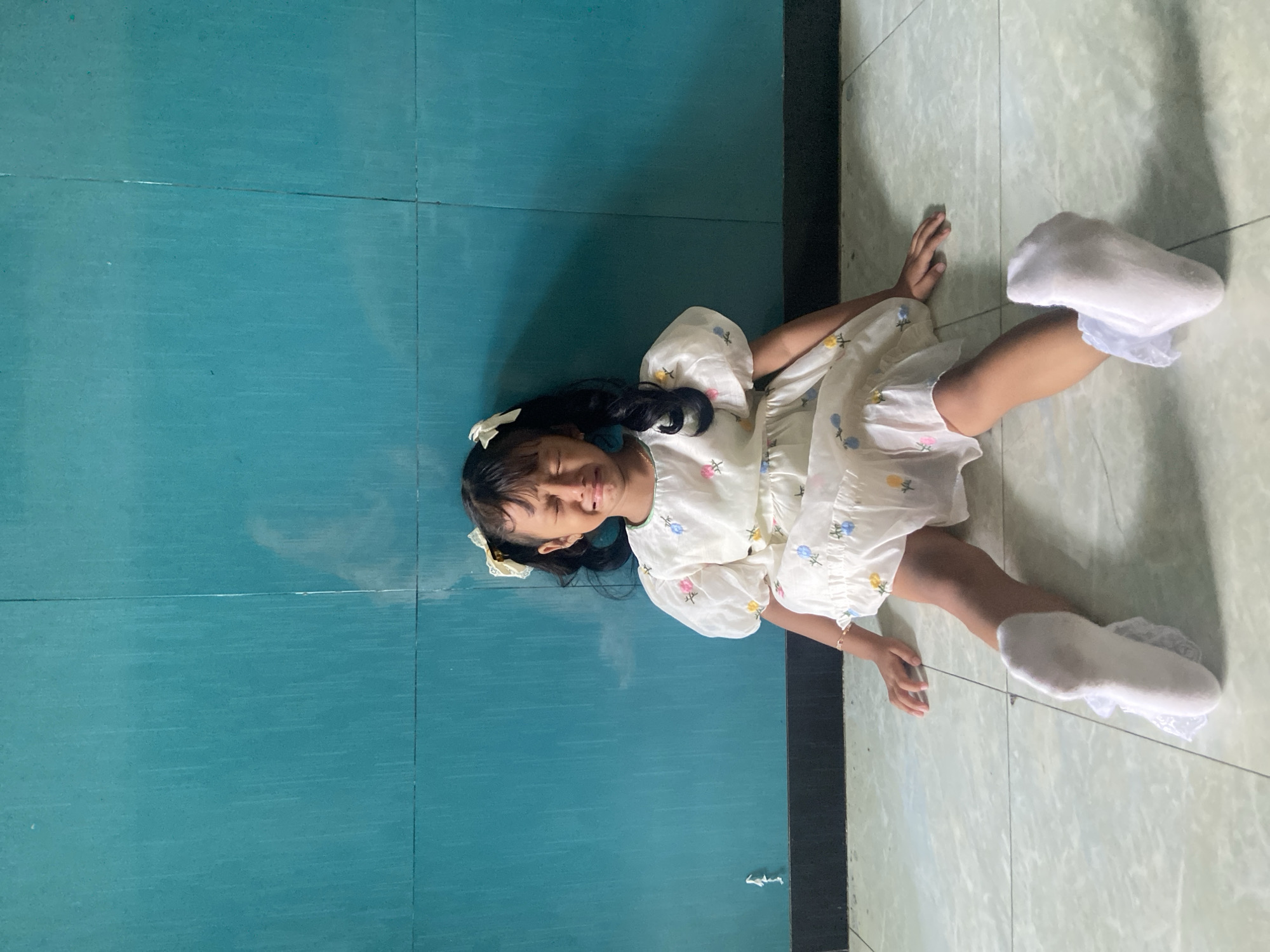 A young girl with dark hair and bangs, wearing a white dress with floral patterns and white socks with lace, lies on the floor. Her head is on a teal tile surface, and her body extends onto a light beige tile surface. The location is Gelam, Indonesia.