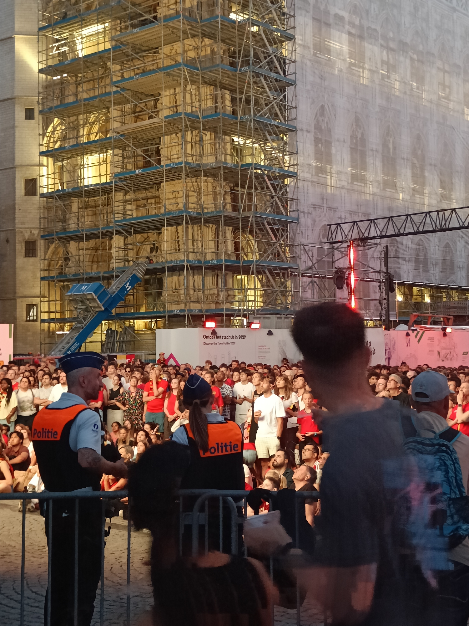 The image depicts an outdoor gathering in front of the Leuven Town Hall, Belgium, which is partially covered by scaffolding and white sheeting, suggesting ongoing renovations. A large crowd of people, predominantly young adults, are assembled, some seated and others standing. Two police officers, identifiable by their "Politie" vests, are positioned in the foreground, maintaining order. The background features the illuminated facade of the town hall, with scaffolding structures extending vertically. In the mid-ground, a large banner with text that reads "Ontdek het stadhuis in 2029 Discover the Town Hall in 2029" is visible, indicating a connection to the town hall's restoration and future accessibility. A stage-like structure with red lighting elements is also present in the background, hinting at a possible event or performance. The overall atmosphere appears to be one of public engagement and anticipation, possibly related to the restoration project or a community event.