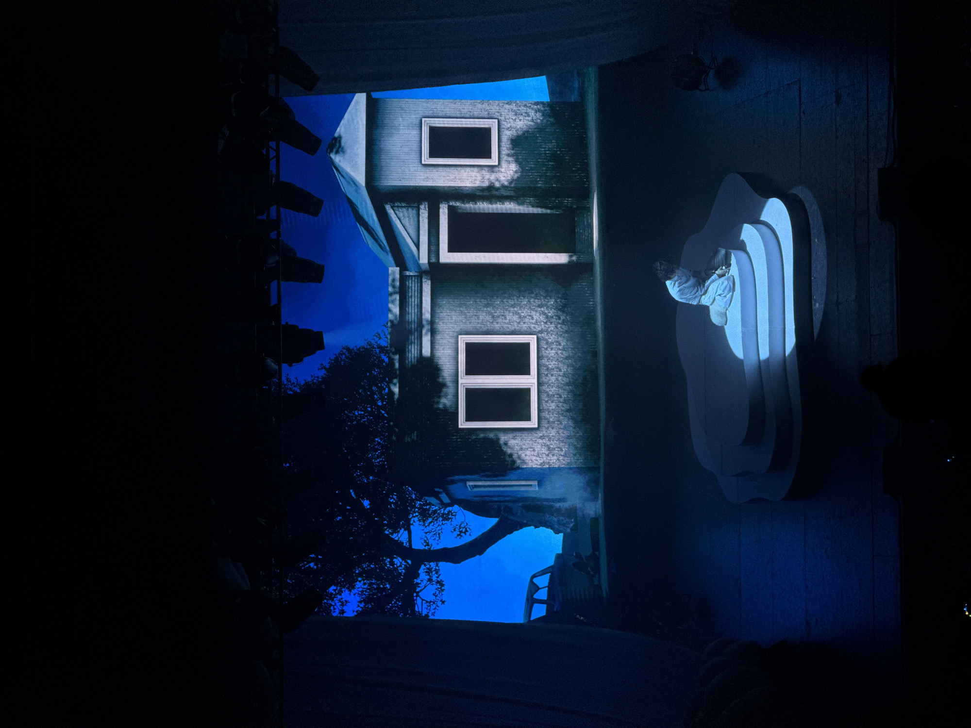 The image captures a theatrical stage set under blue-toned lighting. On the left, there's stage equipment partially visible. The backdrop features a gray, multi-story house with rectangular window frames, set against a blue sky with tree branches visible in the lower section. To the right, a person sits on a multi-tiered, rounded platform, illuminated by a focused light. The overall scene is dark and evokes a sense of drama. Without further details, the location within Los Angeles cannot be determined.