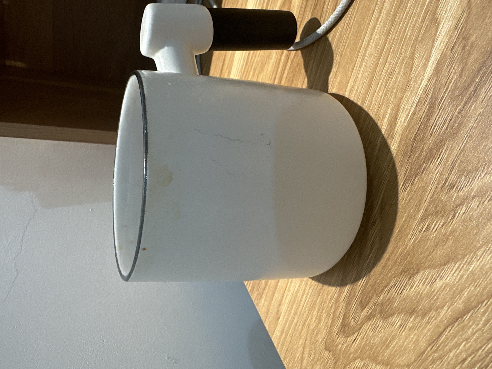 A white cylindrical lamp with a black trim rests on a surface, likely a wooden table or countertop. The lamp has a white cylindrical body and a T-shaped connector at the top, attached to a black cylinder and a white cord. The surface beneath the lamp is a mix of light wood and white material, possibly a wall.