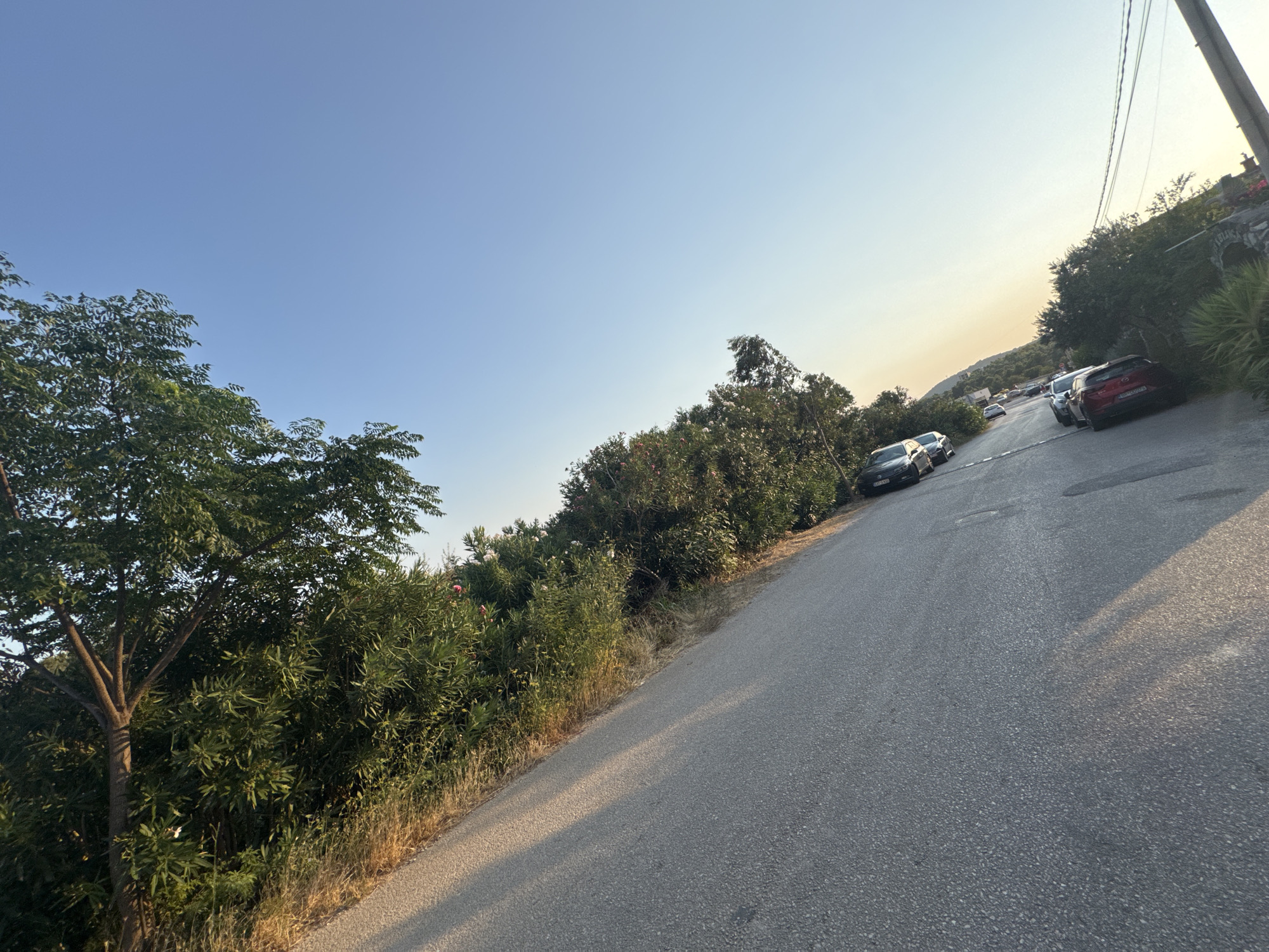 This image captures a sun-drenched, narrow asphalt road winding uphill through a vegetated area in Sutomore, Montenegro. The sky is a clear, pale blue with a gradient towards a pale yellow-orange near the horizon, suggesting either sunrise or sunset. Lush green trees and bushes line the left side of the road, with some dry grass and scrub near the edge. Several cars are parked on the right side of the road, a mix of sedans and SUVs, including a red Mazda CX-5. A utility pole with wires runs diagonally across the upper right portion of the frame. The road is partially shaded by the trees on the left, casting long shadows across the asphalt. In the distance, rolling hills are visible under the soft light. There are no people visible in the frame. The overall scene depicts a quiet, rural or suburban street scene in a Mediterranean climate.