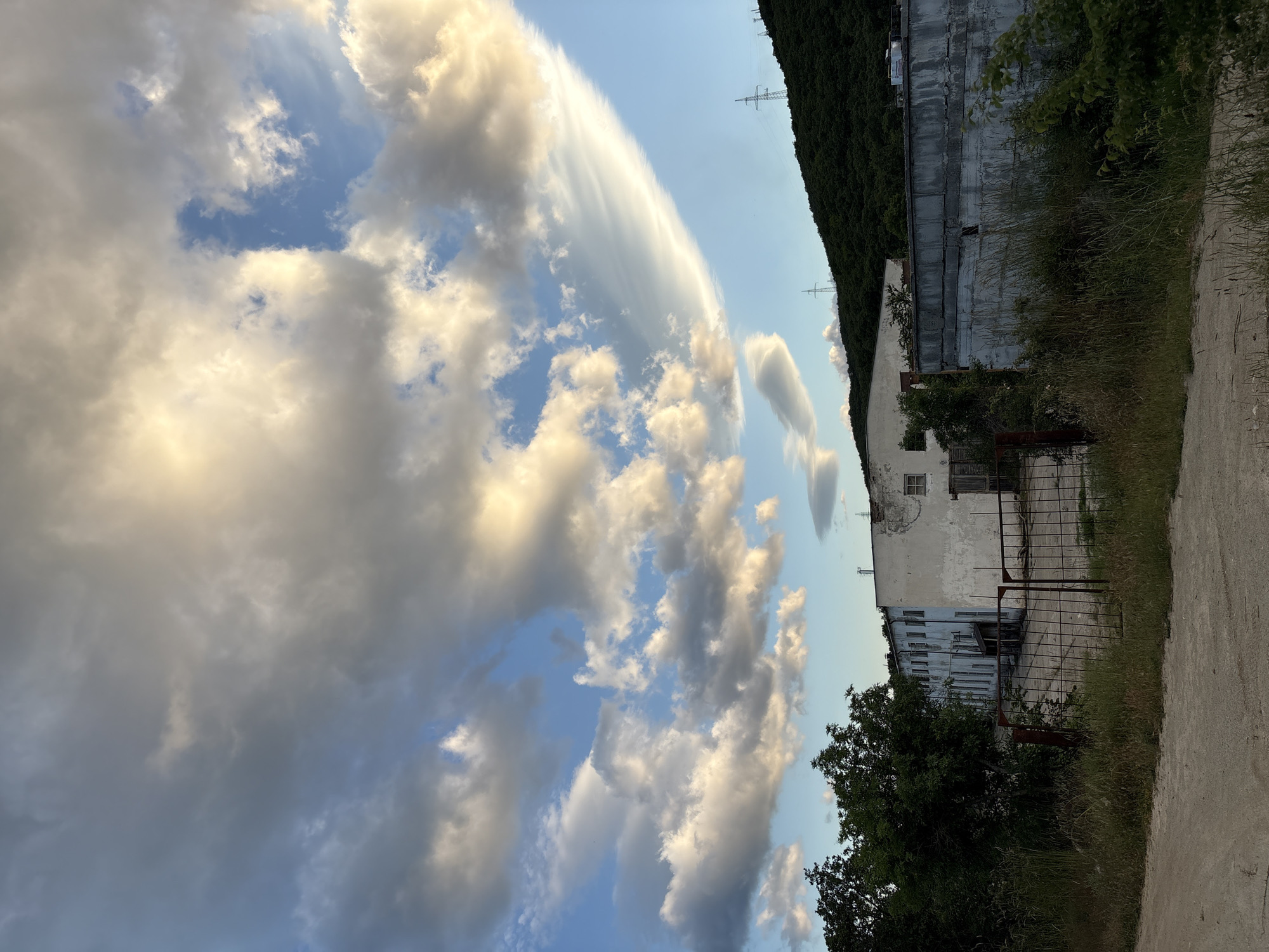 The image depicts an outdoor scene with an industrial building and a lush, tree-covered hillside under a dramatic sky filled with clouds. In the foreground, a dirt path or road leads towards an area with overgrown grass and weeds. A rusty metal gate stands open, revealing a concrete yard in front of a long, white, multi-story building. This building shows signs of disrepair, with peeling paint and some windows boarded up or missing. Adjacent to this building is a lower, concrete structure with a weathered facade.
Behind the buildings, a steep, dark green hillside is densely covered with trees. Several tall electrical pylons are visible on the crest of the hill. The sky above is a striking display of dynamic clouds. Large, fluffy cumulus clouds are illuminated by the setting sun, casting golden and white light, contrasting with areas of blue sky. A distinctive, wispy lenticular cloud stretches horizontally across the upper right portion of the frame. The overall impression is one of an industrial area bordering a natural landscape, captured during the golden hour before sunset.