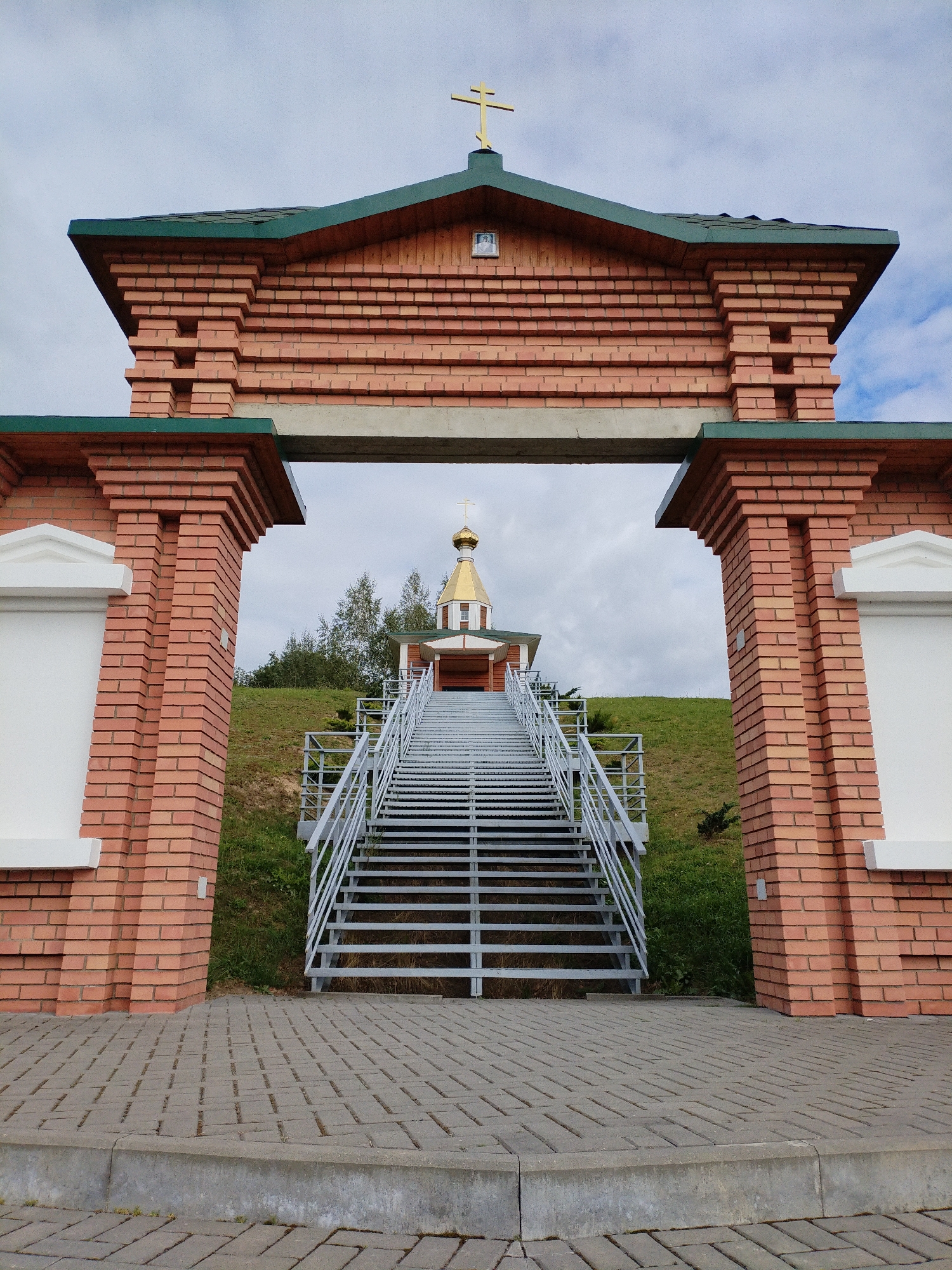 The image captures an outdoor scene featuring an entrance structure with a red brick facade, a green roof, and decorative white elements. A golden cross adorns the top of the structure. Beyond the entrance, a set of metal stairs ascends a grassy hill towards a small building with a golden dome and a cross. The sky is overcast with scattered clouds. The foreground consists of a paved walkway made of gray interlocking pavers. The location is identified as the Chapel of the Holy Protection of the Mother of God in Bialynichy, Belarus. No people or animals are visible in the image.