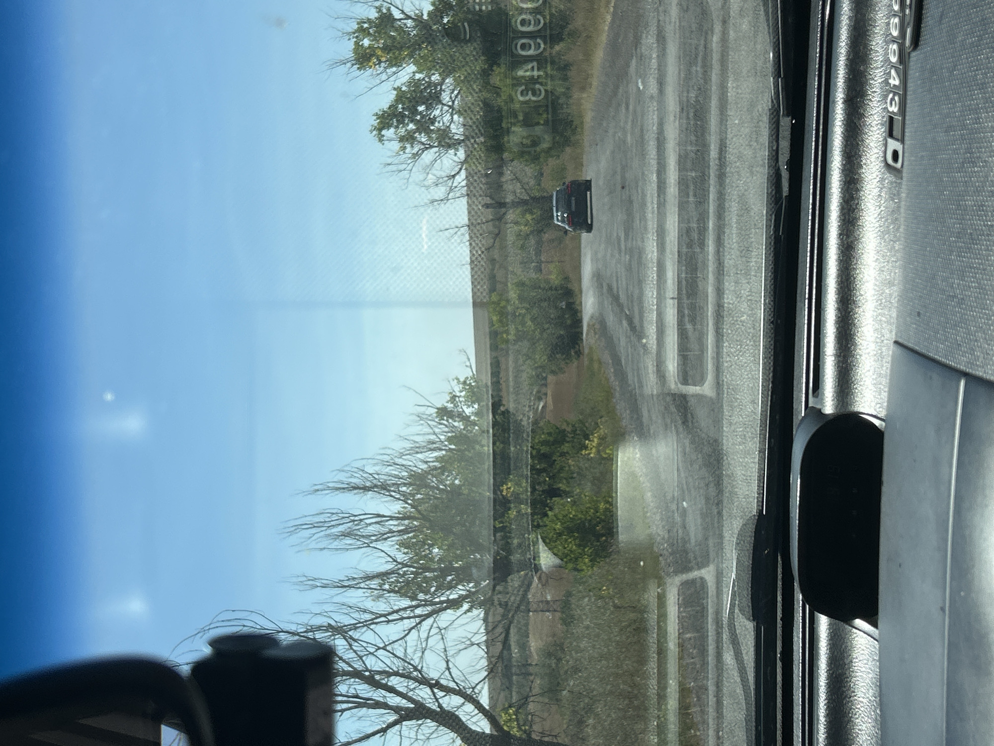 The image, captured from inside a vehicle looking forward through the windshield, depicts a rural road leading towards a slight incline. The sky is a clear blue with two indistinct bright spots. Bare trees and green bushes line the sides of the road. In the distance, a dark-colored car is driving away from the viewer. To the right of the road, a reflective surface with lettering is visible, appearing to be a sign. The dashboard of the vehicle is visible in the foreground, showing a black dash-mounted device and part of a gray textured surface. The presence of a road, vegetation, and a vehicle suggests an outdoor, driving scene. Based on the information provided about the location being Cioc-Maidan, Moldova, this scene likely represents a road within that area.