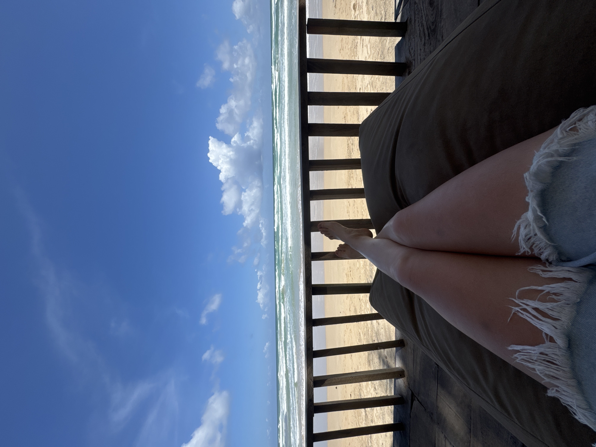 On a sunny day in Ahangama, Sri Lanka, a person sits on a cushioned chair, overlooking a beach. The person's legs, clad in frayed denim shorts, are crossed at the ankles. A wooden railing separates the person from the sandy beach and ocean. Waves crash against the shore. The sky is blue with scattered white clouds.