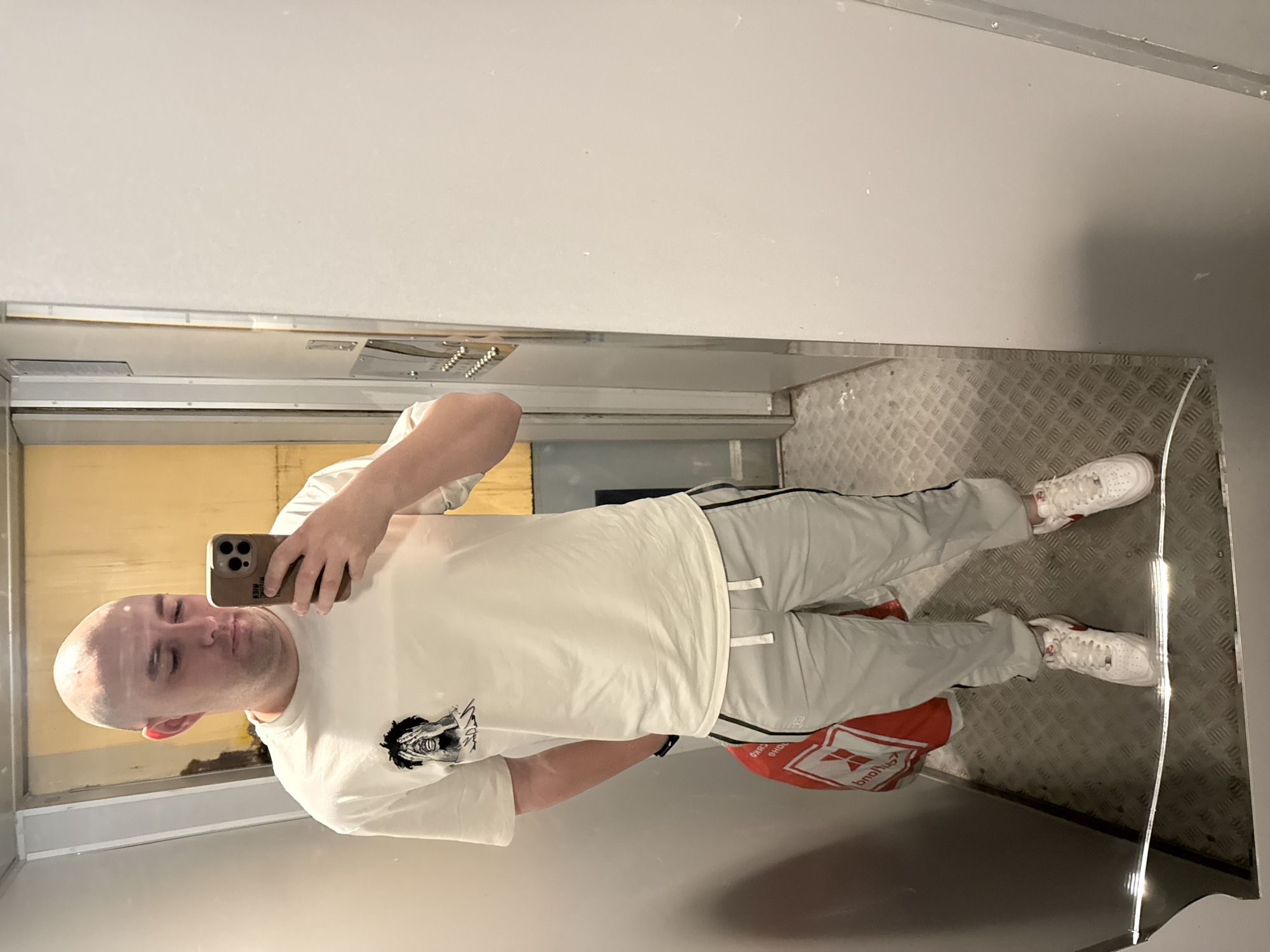 A bald, light-skinned person is taking a selfie in what appears to be a reflective space, possibly an elevator. The person is wearing a white t-shirt with a graphic on the chest, gray sweatpants with a black stripe, and white sneakers with red accents. They are holding a phone with a brown case to take the photo. A red shopping bag is partially visible near their legs. The background consists of reflective metal panels, which create multiple reflections. Based on the information provided the location is in Pleven, Bulgaria. The floor has a diamond plate pattern. The image is tilted to the side.