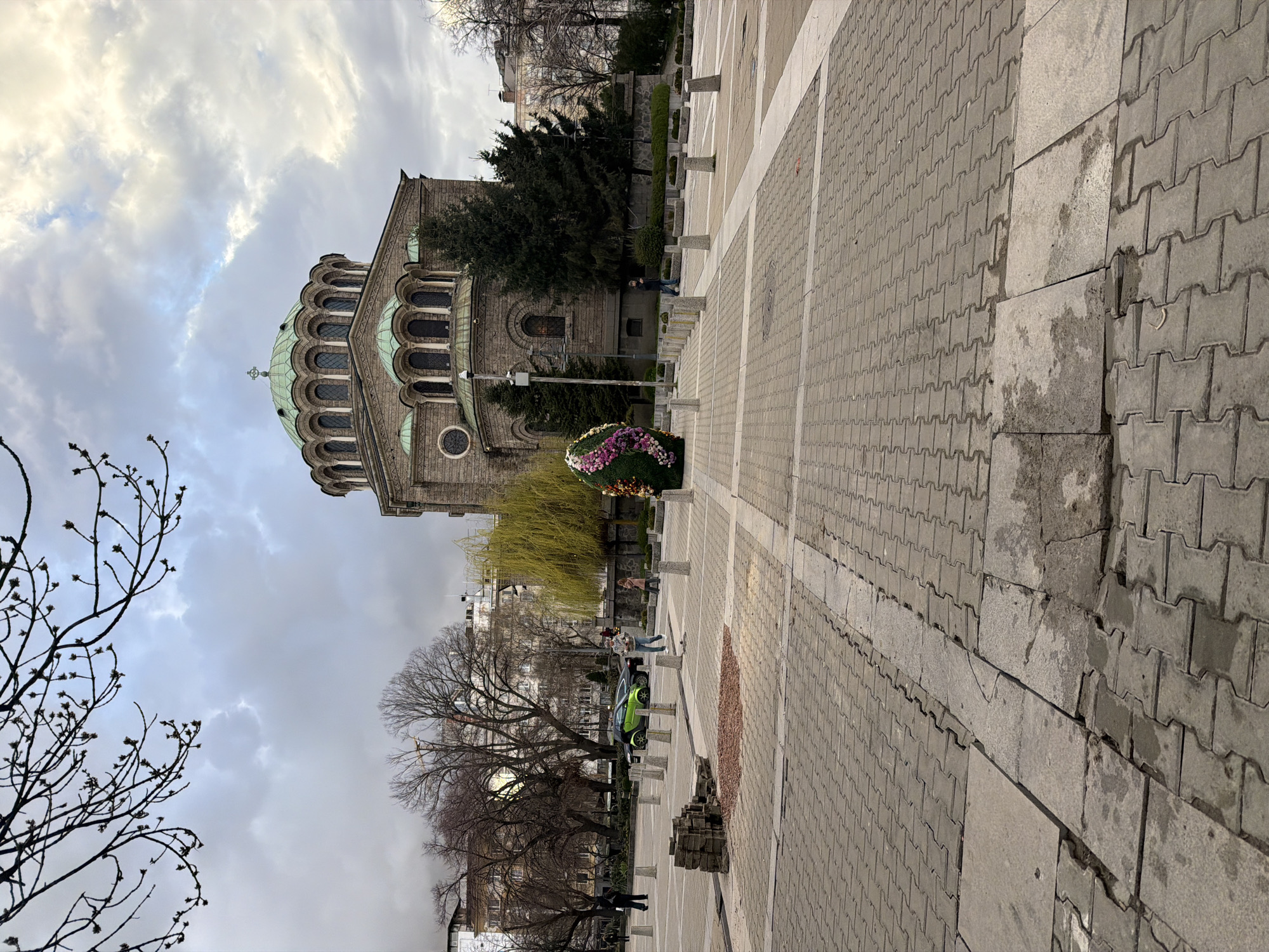 This is an outdoor shot of Alexander Nevsky Square in Sofia, Bulgaria. The Alexander Nevsky Cathedral is visible, featuring a green dome and arched windows. The square is paved with grey blocks. There are sparse trees and leafless branches in the foreground and background. A small green car is visible in the background. A couple of people are walking on the plaza near the cathedral. A wreath-like flower arrangement sits near a tree in the middle distance. The sky is overcast with grey and white clouds.