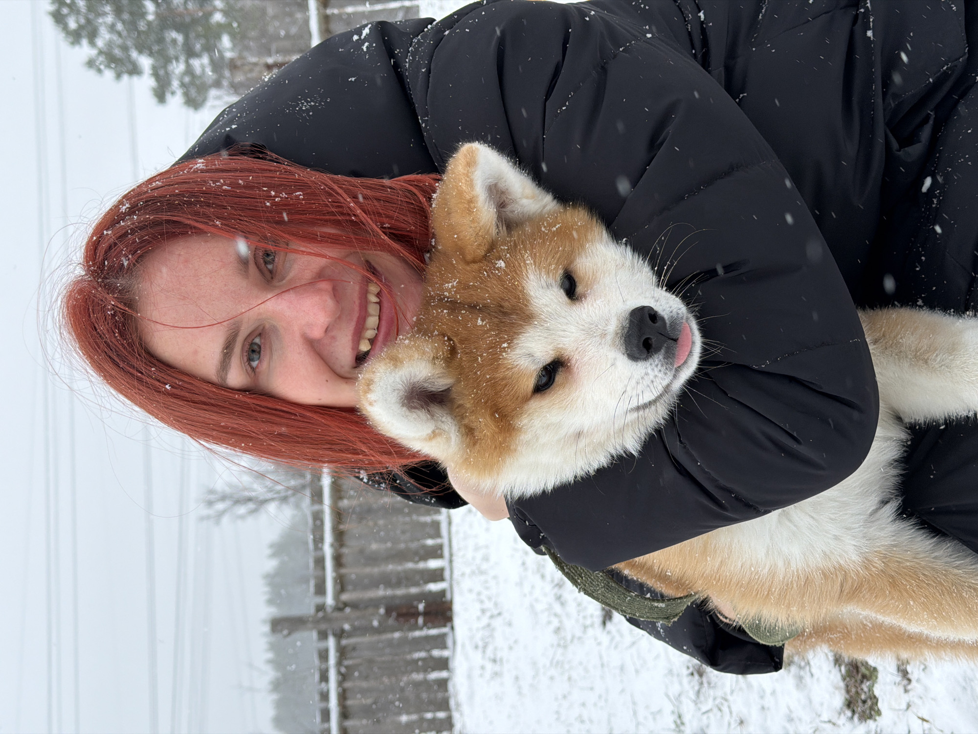 A woman with red hair smiles while holding a brown and white puppy in her arms. Both are outdoors in a snowy environment. In the background are trees and a fence. The scene is likely in Chaikovsky, Russia.