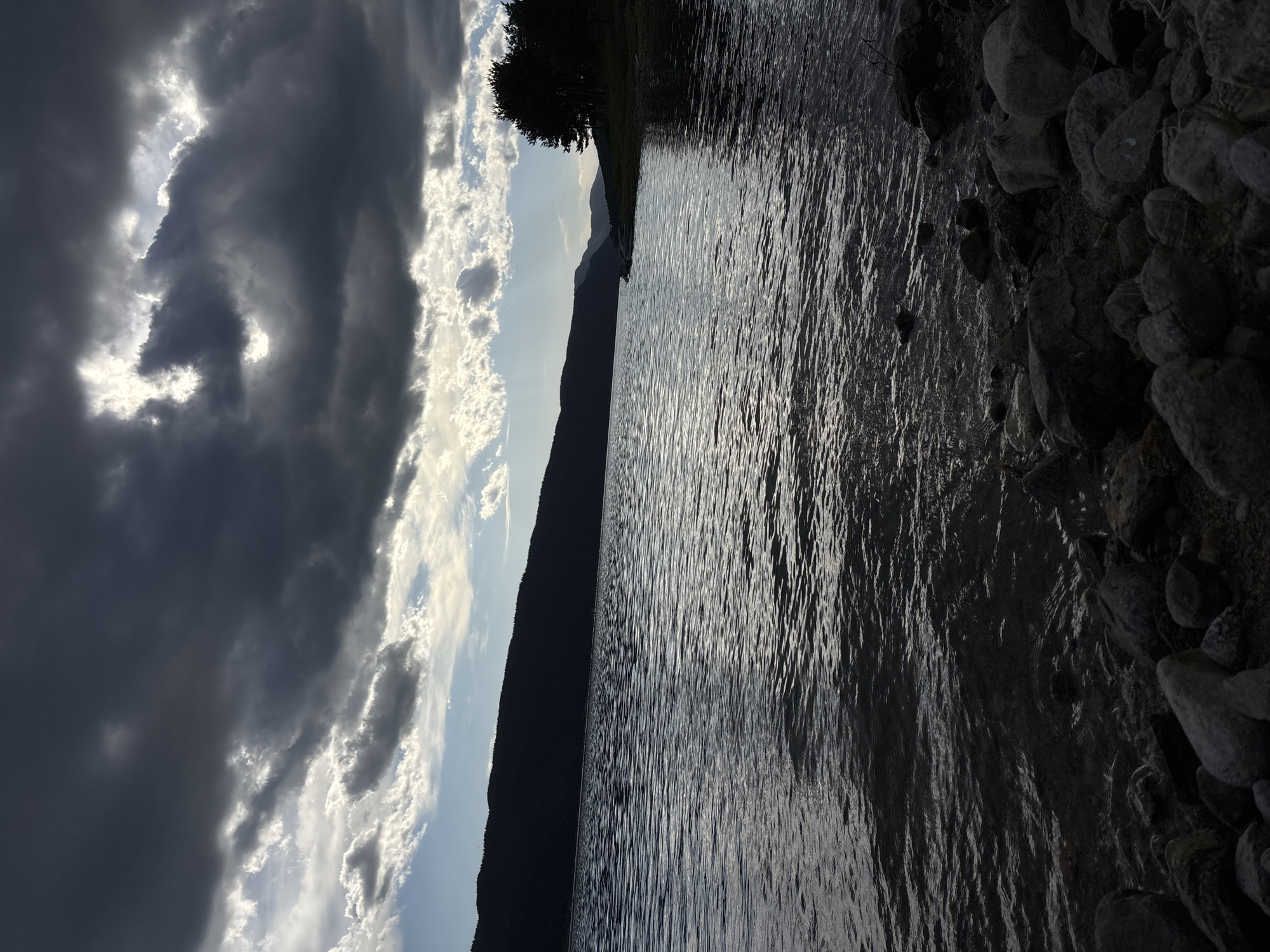 A landscape photograph captures a body of water, possibly a lake or river, with a rocky shoreline on the right and a dark, silhouetted mountain range in the background. The sky is divided between stormy, dark clouds on the left and patches of light blue sky interspersed with white clouds on the right. Reflections of the sky and clouds are visible on the water's surface.