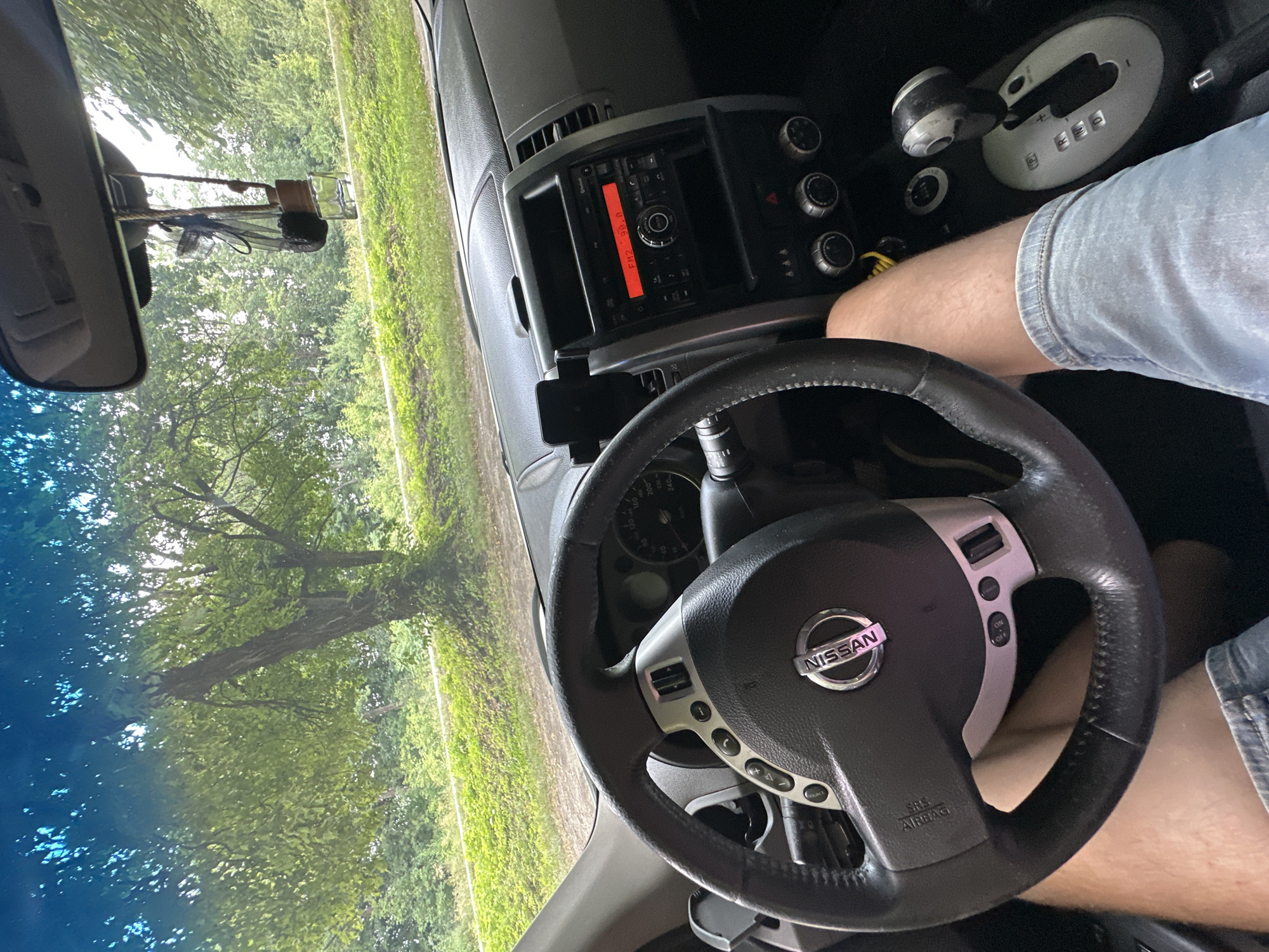 The image is a first-person perspective from inside a Nissan X-Trail vehicle, parked outdoors. The steering wheel, dashboard, and gear shifter are visible. The car's radio is on, displaying "FM2 98.0". Outside the vehicle, a lush green environment with trees and foliage is seen through the windshield, suggesting a rural or park-like setting. The exterior lighting indicates it is daytime. The image was taken in Shatava, Ukraine.
