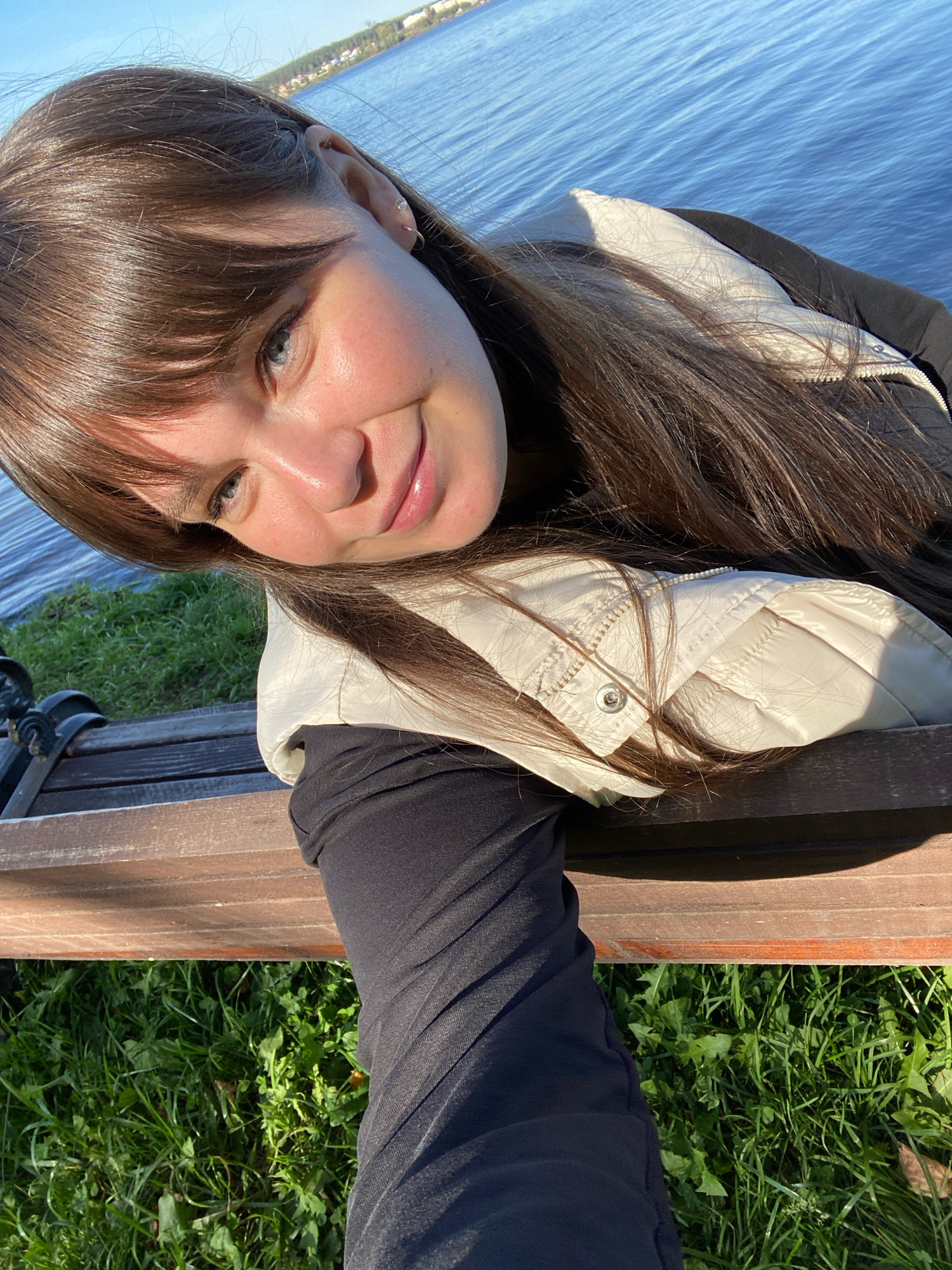 A young woman with long brown hair and a light-colored vest over a black top is captured in a selfie taken outdoors, likely in the city of Votkinsk, Russia. She is seated on a wooden bench next to a body of water, possibly a river or lake, reflecting a clear blue sky. The water is calm with gentle ripples, and the shoreline features lush green grass and vegetation. The bench appears to be made of weathered wood and has a dark metal armrest visible. The woman's facial expression is relaxed and she is smiling faintly. The lighting suggests it is daytime, with sunlight casting shadows.
