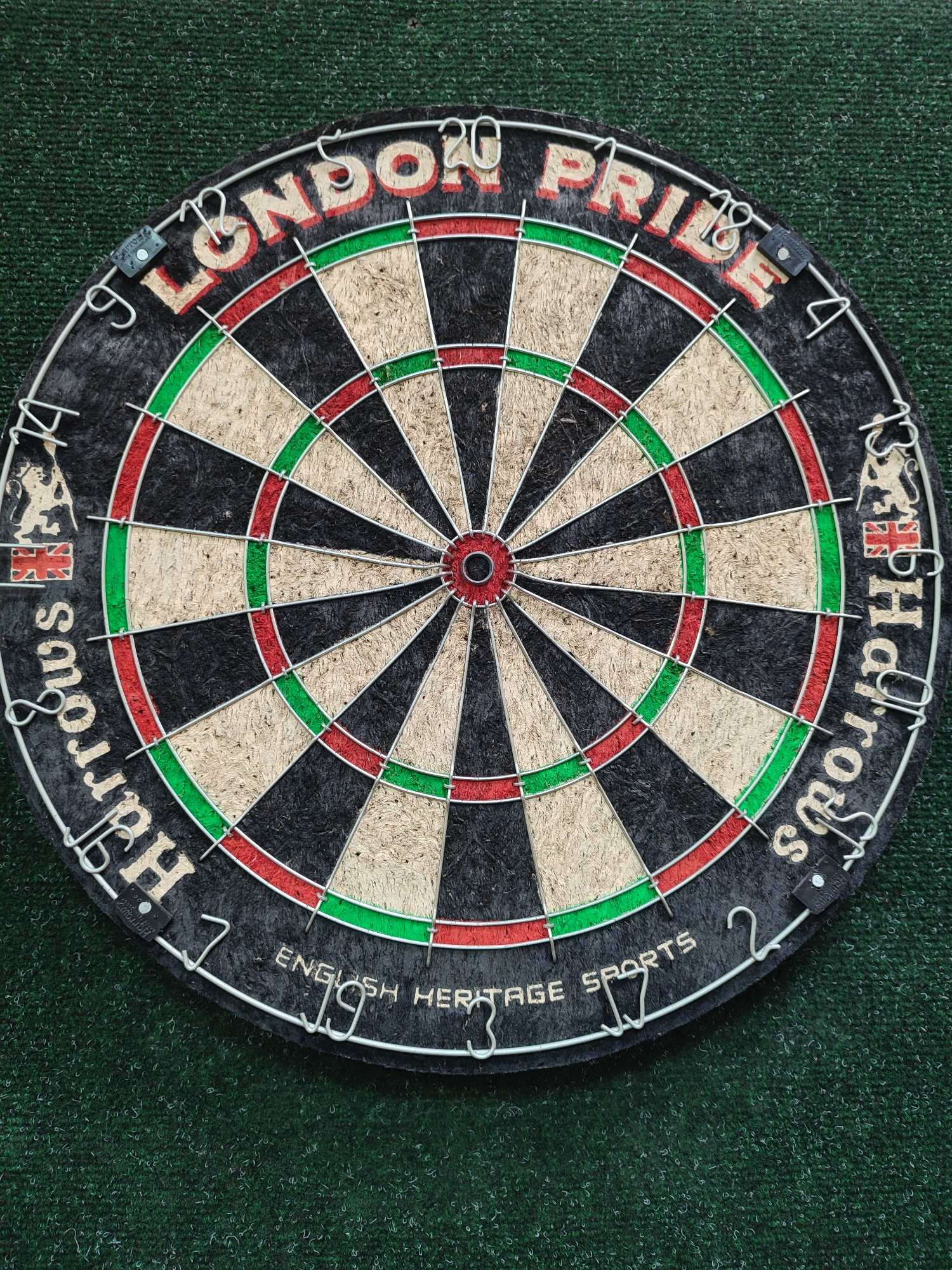 A close-up shot depicts a dartboard featuring "London Pride" and "Harrods" branding, as well as "English Heritage Sports." The dartboard is positioned on a green, carpet-like surface. There is no indication of the location being in Izhevsk, Russia, or any other geographical location. No people, actions, or interactions are visible in the image.