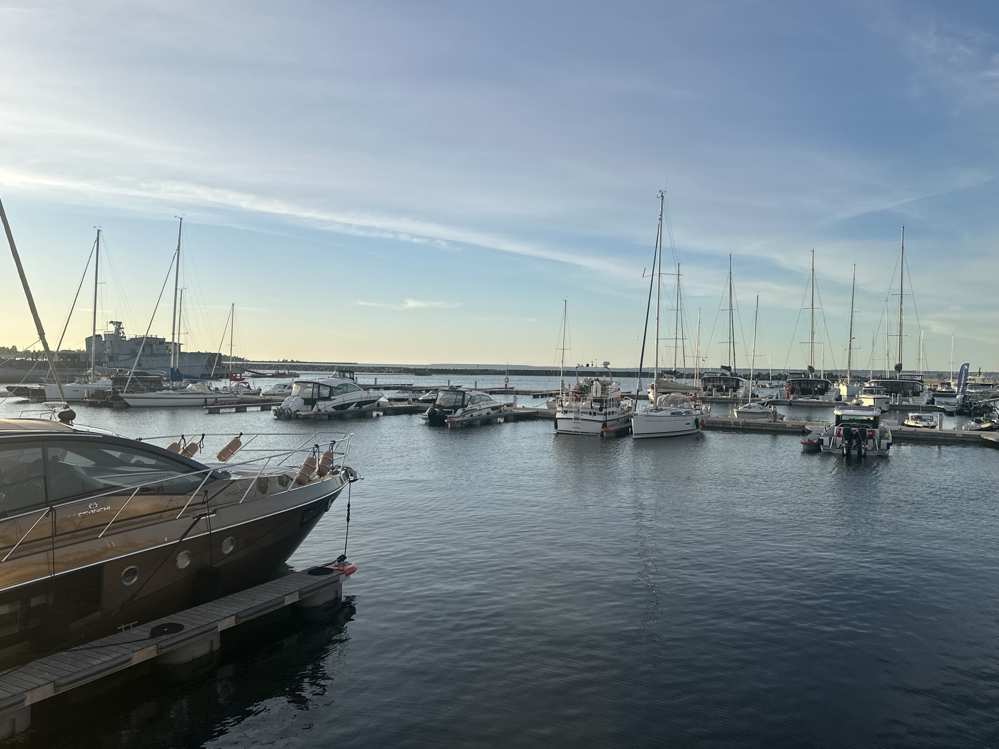 The image shows a harbor in Tallinn, Estonia, with numerous boats docked. In the foreground, a large, sleek, brown yacht is moored to a wooden pier. The yacht has a chrome railing, multiple portholes, and fenders hanging from its side. The water is calm and reflects the sky and the boats. In the background, a variety of sailboats and smaller motorboats are visible, along with a large industrial-looking ship or structure on the left side of the frame. The sky is a clear blue with some wispy clouds, and the overall lighting suggests it is late afternoon or early evening. The scene depicts a tranquil harbor setting with a mix of recreational and possibly commercial vessels.