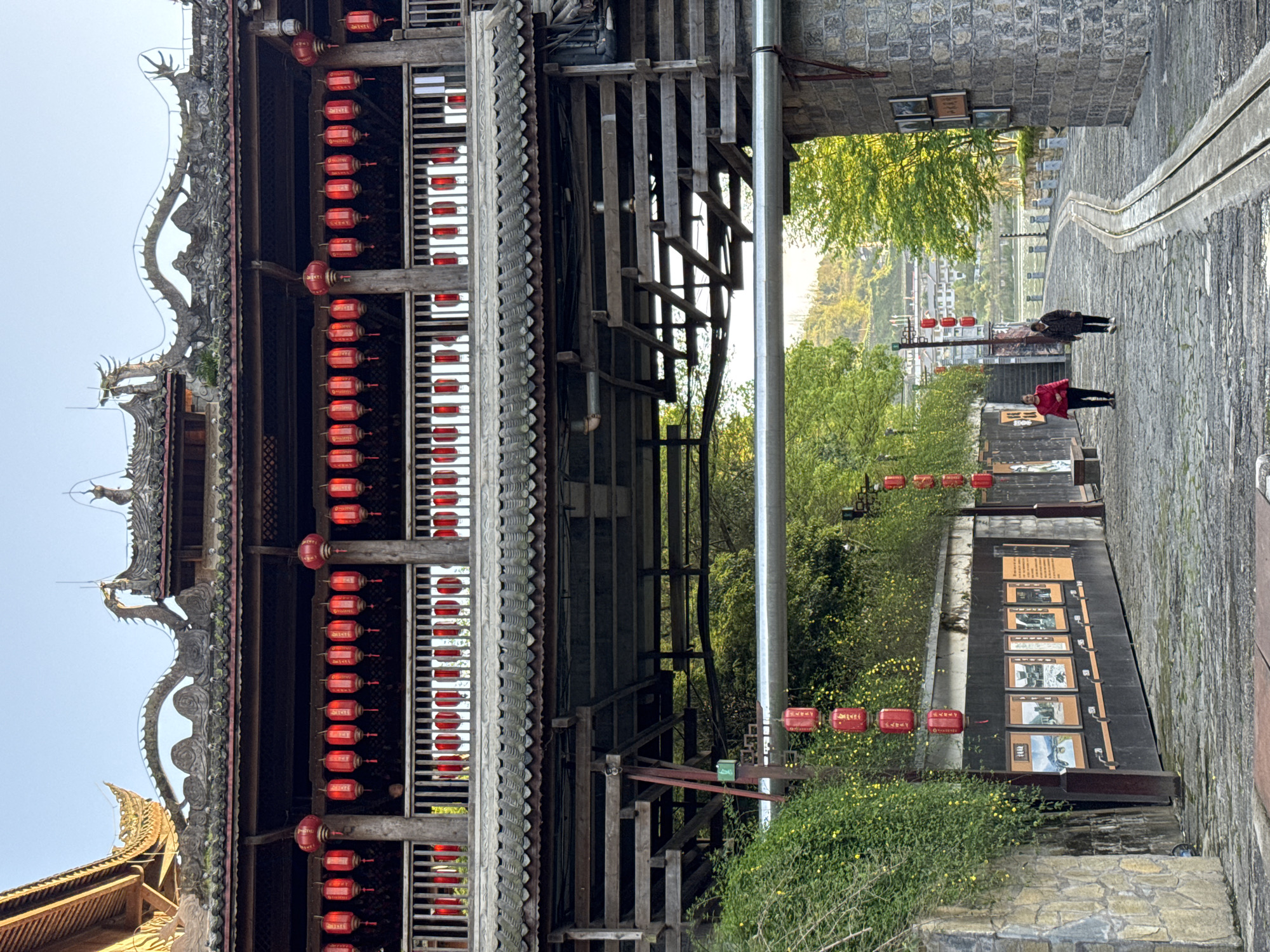 This image shows an outdoor scene in Zhuoshui, China. The foreground features a traditional wooden structure adorned with ornate dragon carvings on its roof and rows of red lanterns. This structure appears to be part of an entrance or gateway leading to a stone-paved street. A large metal pipe runs through the wooden structure, contrasting with the traditional architecture.
Beyond the structure, the street slopes downward and is lined with stone walls. On the right side of the street, a display with informational plaques and images is visible. Further down the street, two people are walking away from the camera. Trees and greenery fill the spaces between the buildings and walls. In the distance, buildings and a body of water are visible under a clear sky.