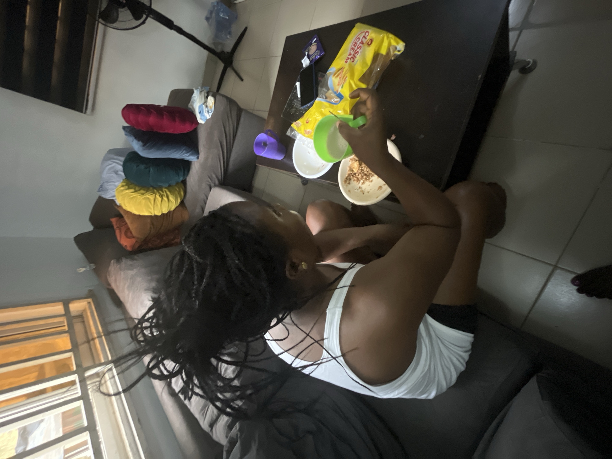 The media file shows a young woman with dark braided hair, wearing a white tank top and black shorts, sitting on a couch. She is holding a green cup and appears to be eating from a bowl placed on a dark coffee table. On the coffee table, there is also a yellow bag of cookies, a purple cup, and a phone. A stack of colorful pillows is visible on the couch beside her. In the background, a fan is mounted on the wall, and a window with blinds is to the left. The scene is indoors, with tiled flooring visible. The lighting suggests it could be daytime or evening. The overall mood appears casual and relaxed. The text "CLASSIC CREAM" is visible on the yellow cookie bag.