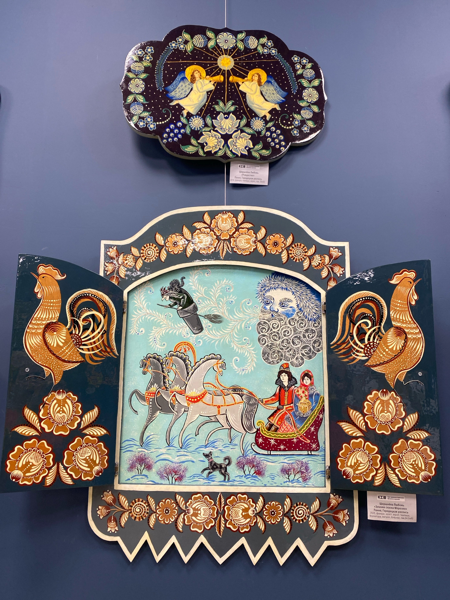 This image depicts a decorative, folk art-style painting displayed on a blue wall, likely in a gallery or exhibition space in Ivanteyevka, Russia, given the exhibition label present. The painting is a hinged, cabinet-like structure with two large side panels resembling doors. Each side panel features a prominent, stylized golden rooster with intricate brown and white feather details, set against a dark teal background. These panels are adorned with decorative floral motifs in shades of brown and cream.
The central part of the painting, visible when the "doors" are open, depicts a winter scene. In the foreground, a horse-drawn sleigh carries two figures, a woman in a red coat and hat holding a basket, and a person next to her wearing a blue coat and red head covering. A small black dog runs alongside the sleigh. In the background, a blue sky with swirling white patterns forms the backdrop. A stylized face with a blue beard and hair, resembling a winter spirit, is depicted in the upper right portion of the central panel. A witch on a broomstick flies across the top of the scene. The entire central panel is framed by decorative floral patterns in orange and white on a light blue background, and a darker teal border at the top and bottom. The bottom of the structure is cut into a decorative, sawtooth pattern.
Above this main artwork, a smaller, dark blue, scalloped plaque is also displayed. This plaque features a central image of an angel with blue wings blowing a golden trumpet, surrounded by stars and stylized floral elements in blue, white, and yellow. A small label is attached to the bottom edge of the main artwork, providing exhibition details.