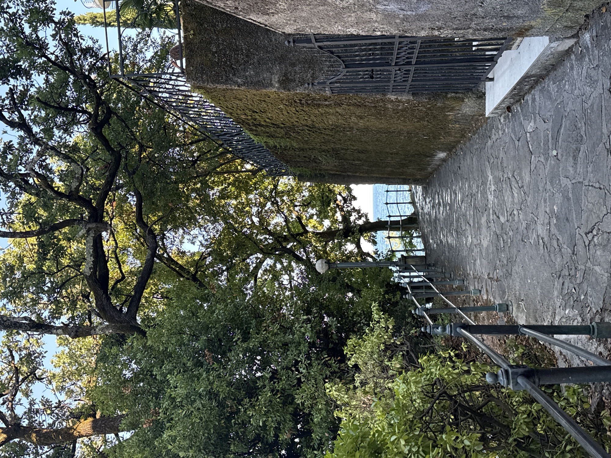 A stone pathway slopes gently downwards, bordered by lush green foliage on the left and a moss-covered stone wall topped with an ornate black metal fence on the right. The pathway is paved with irregularly shaped grey stones. A dark metal railing with decorative finials runs along the left side of the path. Sunlight filters through the leaves of large trees overhead, casting dappled shadows on the ground. In the distance, a body of water, likely the Adriatic Sea, is visible beyond a black metal fence and a single lamppost. The scene appears to be located in Grad Opatija, Croatia, given the architectural style and vegetation commonly found in coastal Adriatic towns.