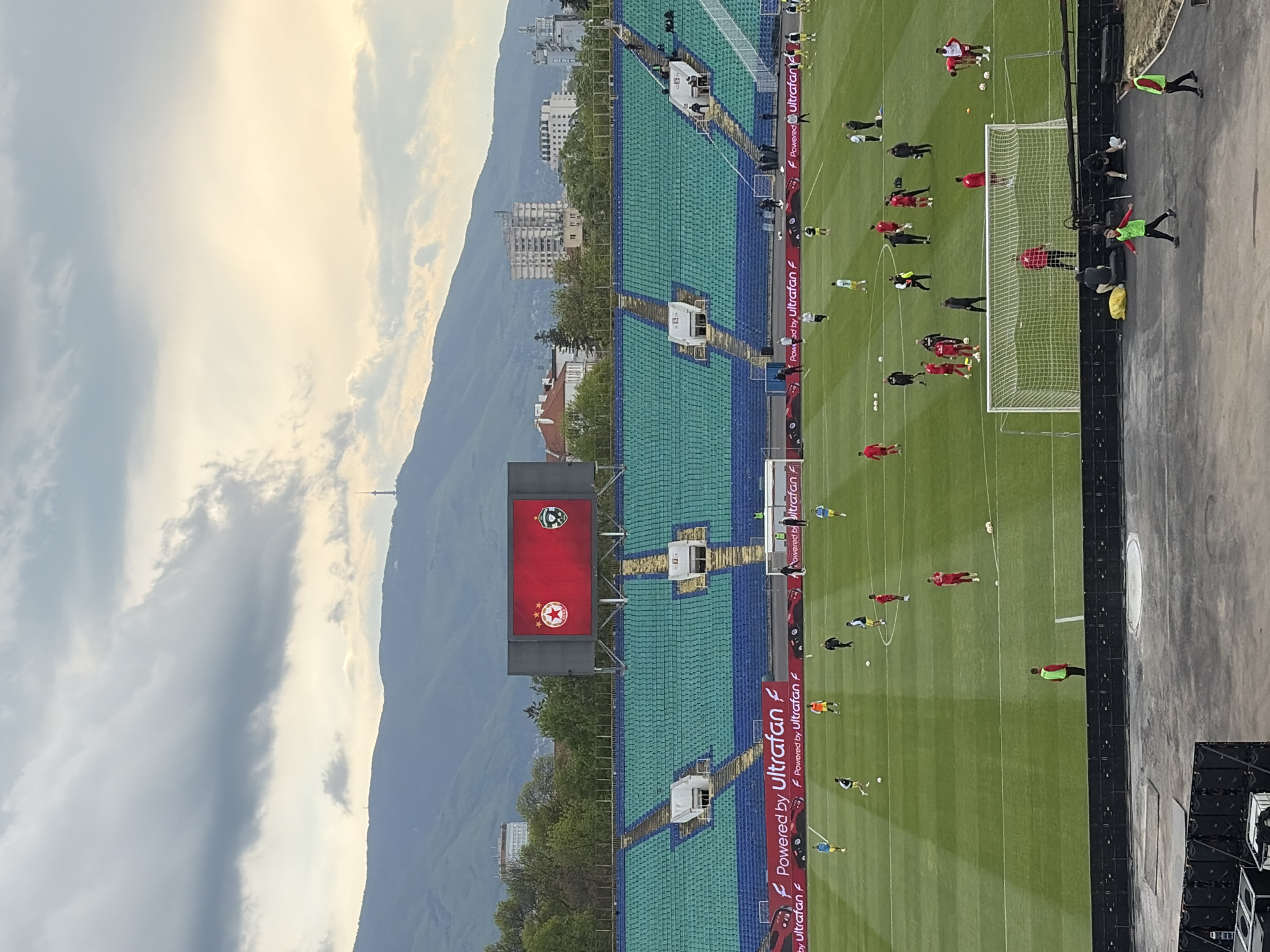 This image, rotated 90 degrees clockwise from its intended upright orientation, captures a football stadium scene, likely in Sofia, Bulgaria, given the distinctive mountainous backdrop. The photograph appears to be taken in the late afternoon or early evening under a cloudy sky, with soft light illuminating the scene.
The central focus is a vibrant green football pitch where numerous players are engaged in a warm-up or practice session. Many players are dressed in red kits, while others wear yellow, light blue/green, or black uniforms. They are spread across the field, with several players near a white goalpost on the right side of the frame. Coaches or staff members are also visible, some wearing high-visibility green and yellow vests.
The stadium features extensive seating areas in alternating patterns of blue and green, suggesting a large capacity venue. Elevated structures, possibly for media or security, are integrated into the stands. A prominent red scoreboard on the left side displays two distinct football club logos: one red with a white star, and another green.
Along the perimeter of the pitch, advertising hoardings repeatedly display the text "Powered by Ultrafan F" and variations thereof. Beyond the stadium, a glimpse of urban buildings transitions into the imposing, dark silhouette of mountains dominating the horizon, partially veiled by clouds. A faint tower can be seen atop one of the distant peaks. The overall impression is one of anticipation or preparation within a sporting venue nestled against a grand natural landscape.
