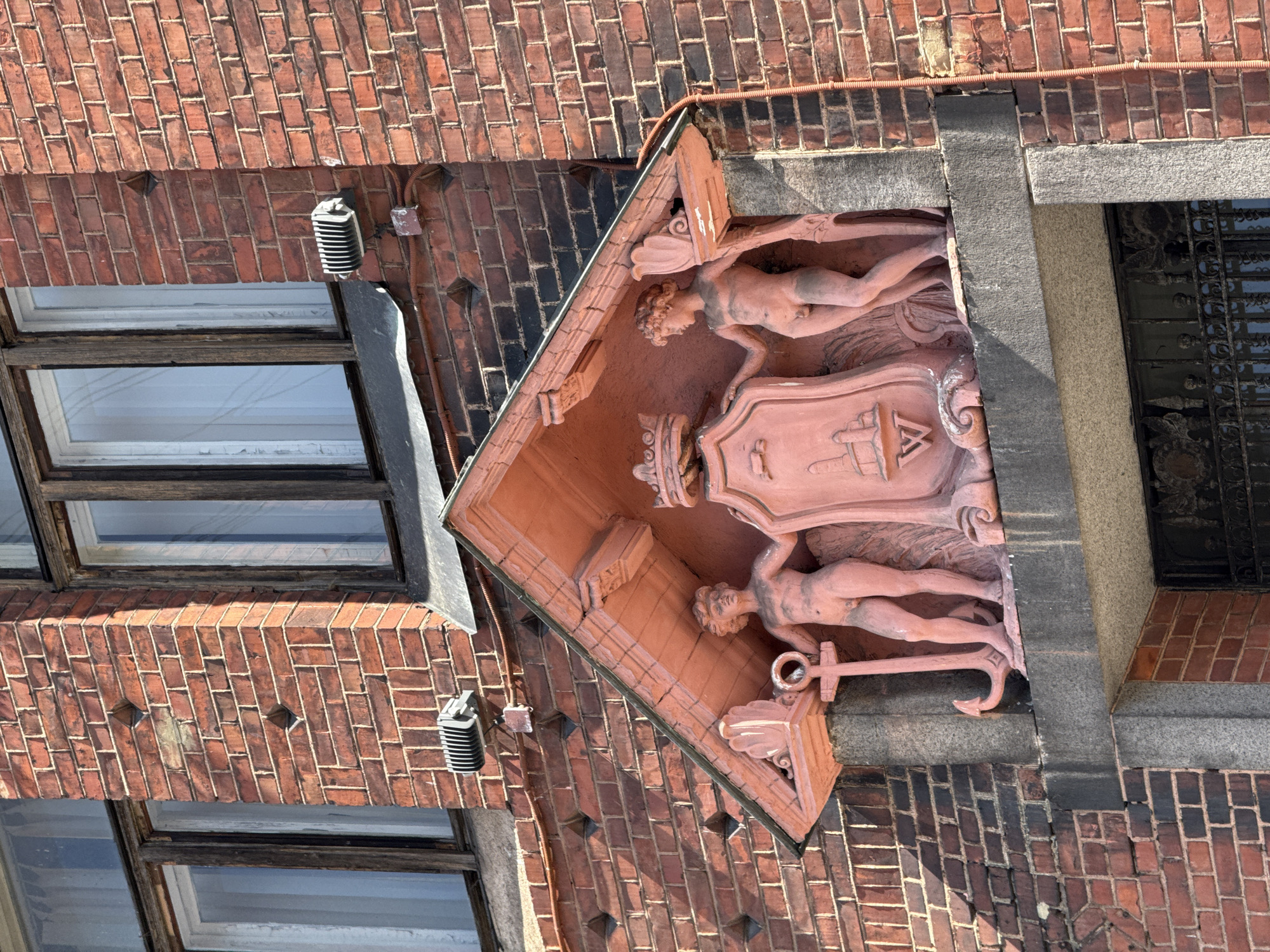 The image depicts an architectural detail on a red brick building. A salmon-pink colored relief adorns the facade, situated beneath a pitched roof-like structure. The relief features two nude figures of boys, one on the left holding an anchor and the other on the right. Between them, a crest with a crown on top is displayed, along with an image of buildings and a stylized letter "W". The figures are placed on a stone ledge. Above the relief, two modern grey light fixtures with ribbed covers are mounted on the brick wall, connected by orange electrical conduit. The brickwork shows a diamond pattern in some sections. Two windows with white curtains are visible on the upper part of the building. This architectural element is located on the facade of the Vyborg State Technical College building, formerly known as the Vyborg Trade School, located at 6, Vyborgskaya Street, Vyborg, Russia.