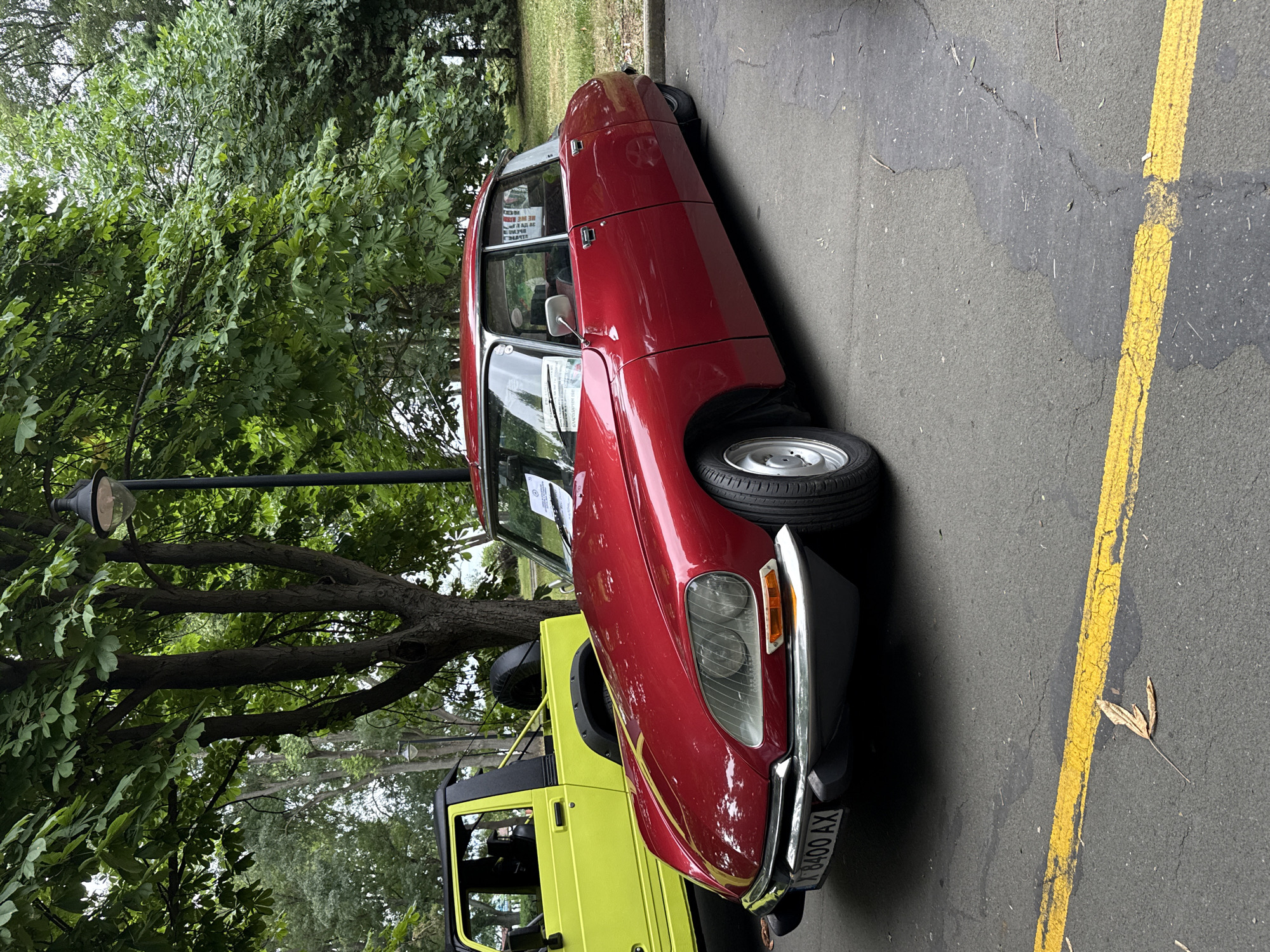 The image shows two vehicles parked on an asphalt surface with a yellow line marking. A red vintage Citroën DS is parked on the right, and a bright yellow off-road vehicle is on the left. The red car has a license plate that reads "X 8400 AX". The background features trees and foliage, suggesting an outdoor setting. The scene appears to be part of a car show or event. The location is specified as the city of Burgas, Bulgaria. There are no people visible in the image. The environment is temperate and appears to be daytime, with overcast sky.