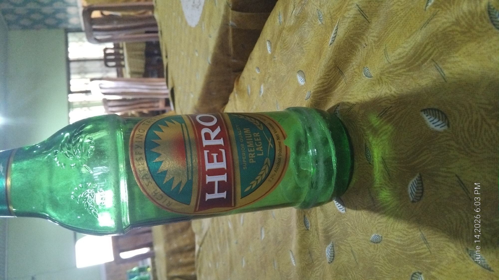 An extreme close-up, vertical shot shows a green glass beer bottle on a yellow tablecloth with a repeating leaf pattern. The bottle is upright, and the label is partially visible, displaying the word "HERO" in large white letters within a red and gold design. Above "HERO," the text "RICH SATISFYING TASTE" is visible in a circular pattern, and below it, "SUPERIOR QUALITY PREMIUM LAGER" is printed. The bottle has embossed detailing near the neck. The lighting suggests an indoor setting, possibly a restaurant or bar, with diffused light creating a shadow of the bottle on the tablecloth. The background is blurred, showing wooden chairs and tables, hinting at a dining area. The timestamp "June 14, 2026 6:03 PM" is visible in the bottom left corner. The location of Howayi, Nigeria is not depicted or inferable from the image content.