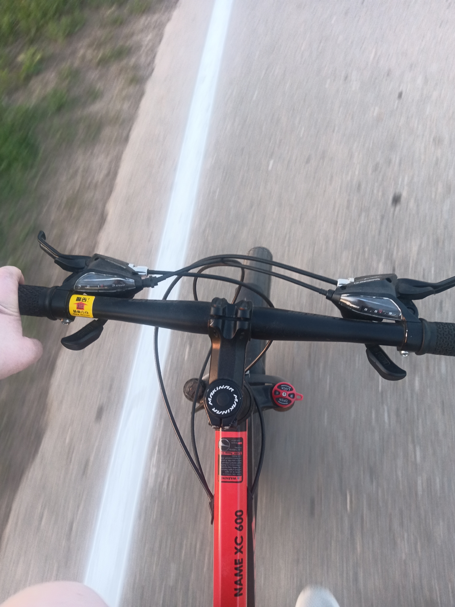 A first-person view from a bicycle moving on a paved road. The bicycle has black handlebars, a red frame labeled "NAME XC 600", and gear shifters. There is a white line on the left side of the road. A blurry patch of grass is visible on the far left. The location is unknown and cannot be determined from the image. No people are fully visible, only a partial hand and leg. The bicycle is in motion.