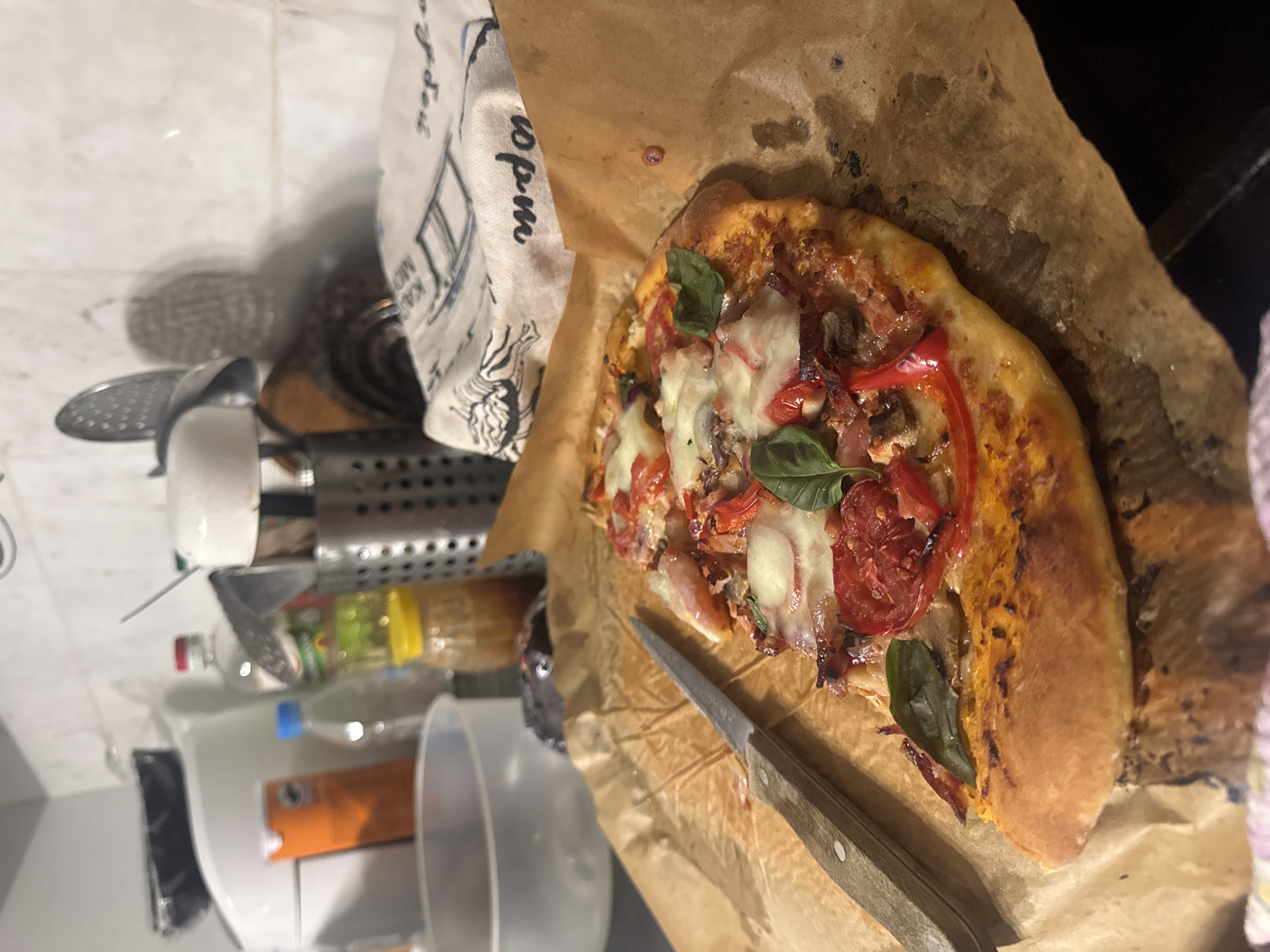 A freshly baked homemade pizza is the central focus of this indoor kitchen scene, presented on crumpled brown parchment paper. The pizza is generously topped with melted mozzarella cheese, sliced red tomatoes, basil leaves, red bell peppers, and what appear to be mushrooms and possibly onions. Its crust is golden brown, indicating it's just out of the oven. A kitchen knife with a wooden handle rests alongside the pizza, suggesting it is about to be cut and served.
To the left of the pizza, a white kitchen towel with blue illustrations and text is visible. Partial text like "opm" and "KAA MIO" can be discerned on the towel. The background reveals a cluttered kitchen environment, typical of a home setting. Various kitchen utensils and containers are visible, including a metal utensil holder, white mugs, a metal strainer, and several plastic bottles containing liquids, possibly cooking oil or water. A clear plastic measuring cup and a larger white plastic bucket are also present. The overall impression is that of a casual home kitchen, likely in the evening given the artificial lighting and the warm glow on the pizza. No people are visible.