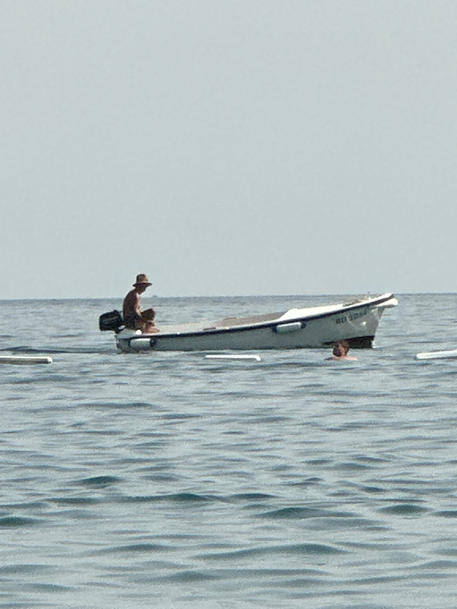 In the city of Boreti, Montenegro, a lone individual wearing a hat is seated in a small white boat, which is anchored in calm, greyish-blue water. The boat has a black outboard motor attached to its stern and appears to have some writing on its side, possibly a registration number. In the water near the stern of the boat, another person's head is visible, suggesting they are swimming or treading water. The background consists of a hazy, light grey sky meeting the horizon line of the water. The overall scene depicts a tranquil day at sea.