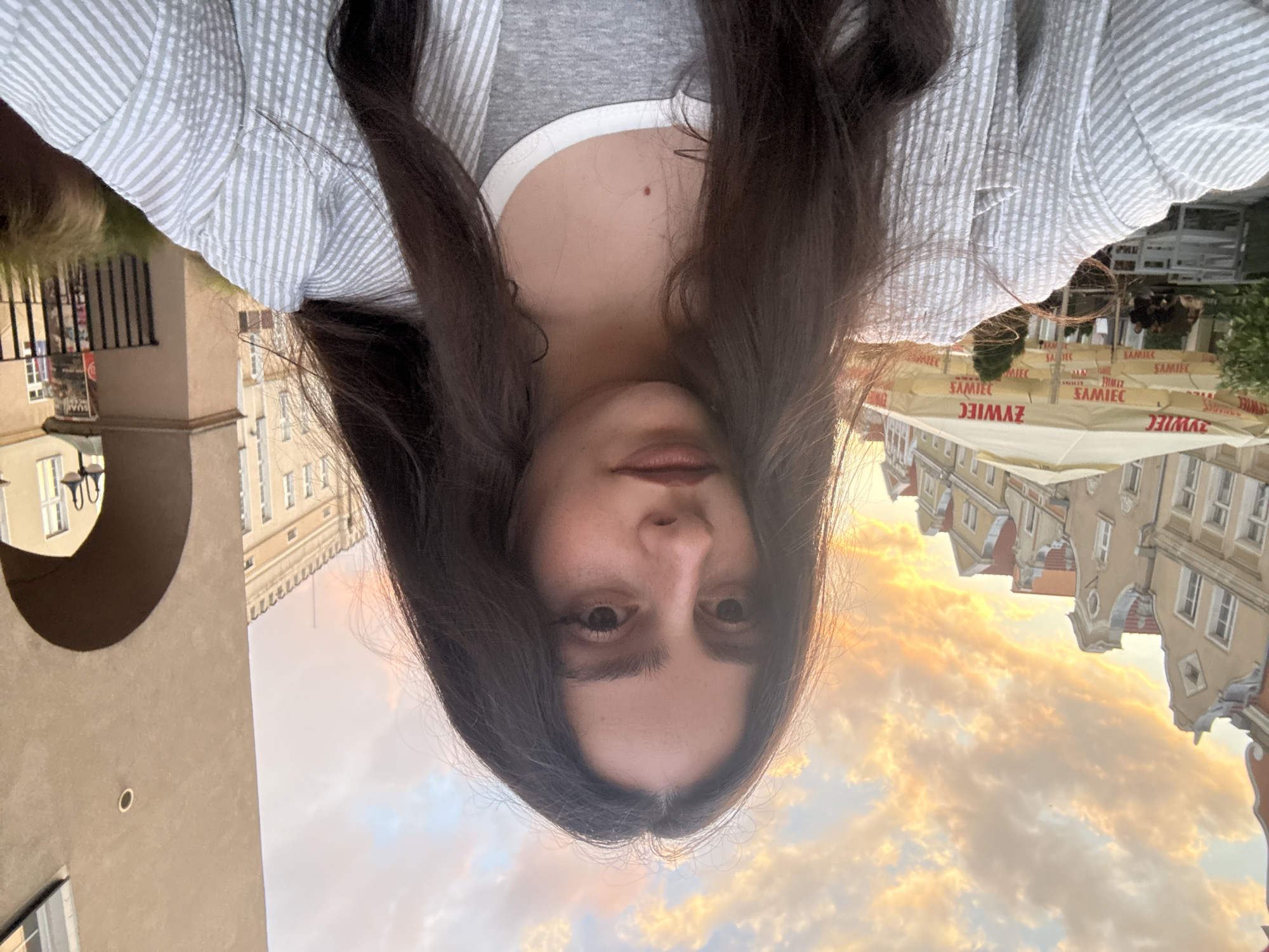 The image is a portrait-oriented selfie featuring a person with long, dark hair looking directly at the camera. They are wearing a light grey striped collared shirt over a plain grey t-shirt. A small mole is visible on their upper chest.
The background reveals an urban setting under a warm, golden sky, indicating late afternoon or early evening during the golden hour. The sky is partially filled with soft, orange and pink-hued clouds. On the right side of the frame, several buildings with distinctive European architecture, including colorful facades and gabled roofs, are visible. Below these buildings, multiple yellow awnings are deployed, clearly displaying the text "ZYWIEC," a Polish beer brand, suggesting an outdoor cafe or restaurant. On the left, a large, light-colored building with an arched opening adds to the architectural variety. The overall scene suggests a pleasant atmosphere in an old town or city square.
Visible text: "ZYWIEC" on the yellow awnings.
The setting is Opole, Poland.