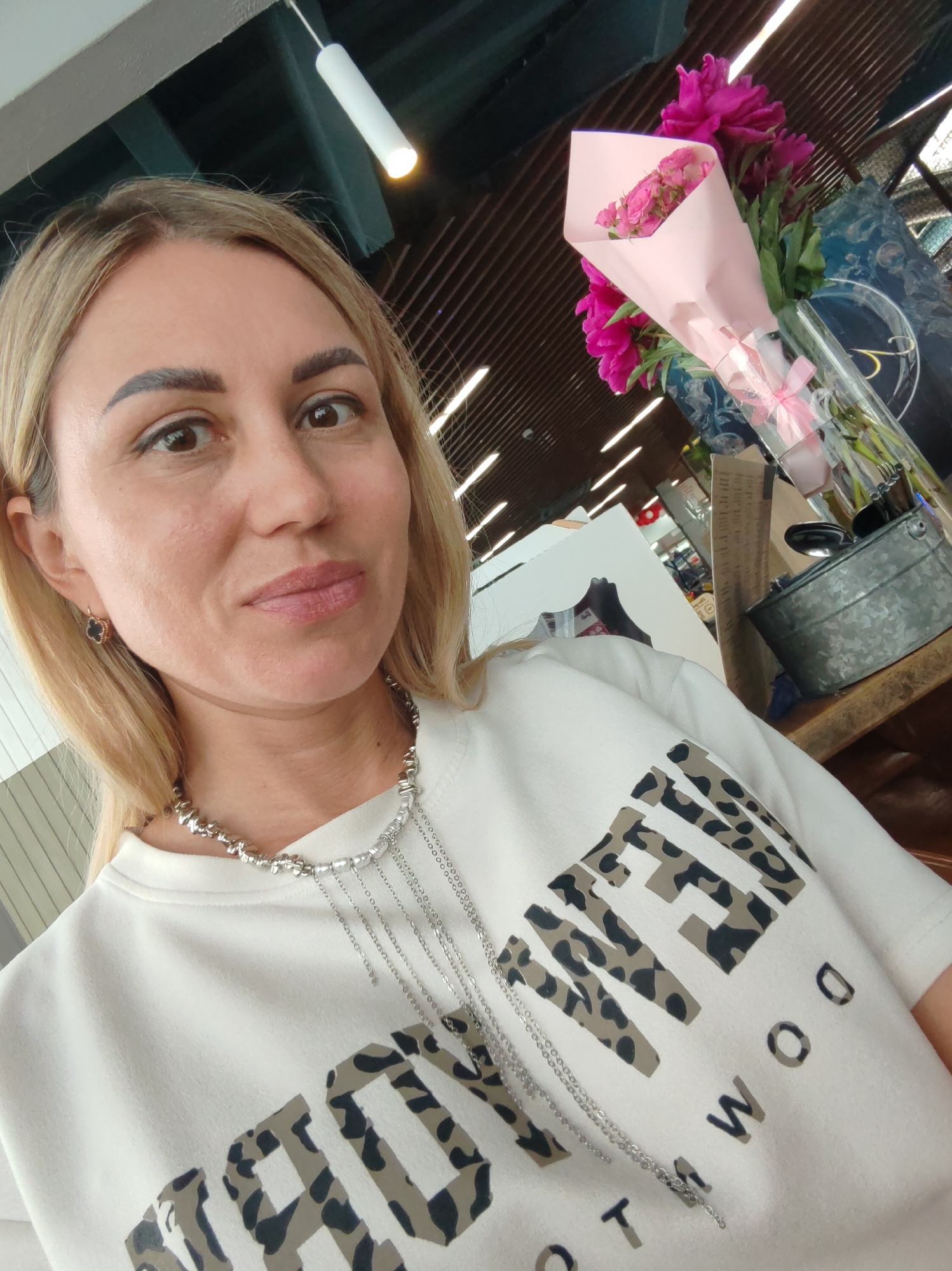 A woman with blonde hair and brown eyes is taking a selfie indoors. She is wearing a white t-shirt with a leopard print pattern on it, and a silver necklace. She has on an earring in her left ear. Behind her, there is a bouquet of pink flowers in a vase, and a wooden table with various items on it, including a metal bucket. The background appears to be a commercial or public space with a ceiling made of wooden slats and bright lighting fixtures. The image is taken from a close-up perspective, focusing on the woman's face and upper body. The location is identified as Amursk, Russia.