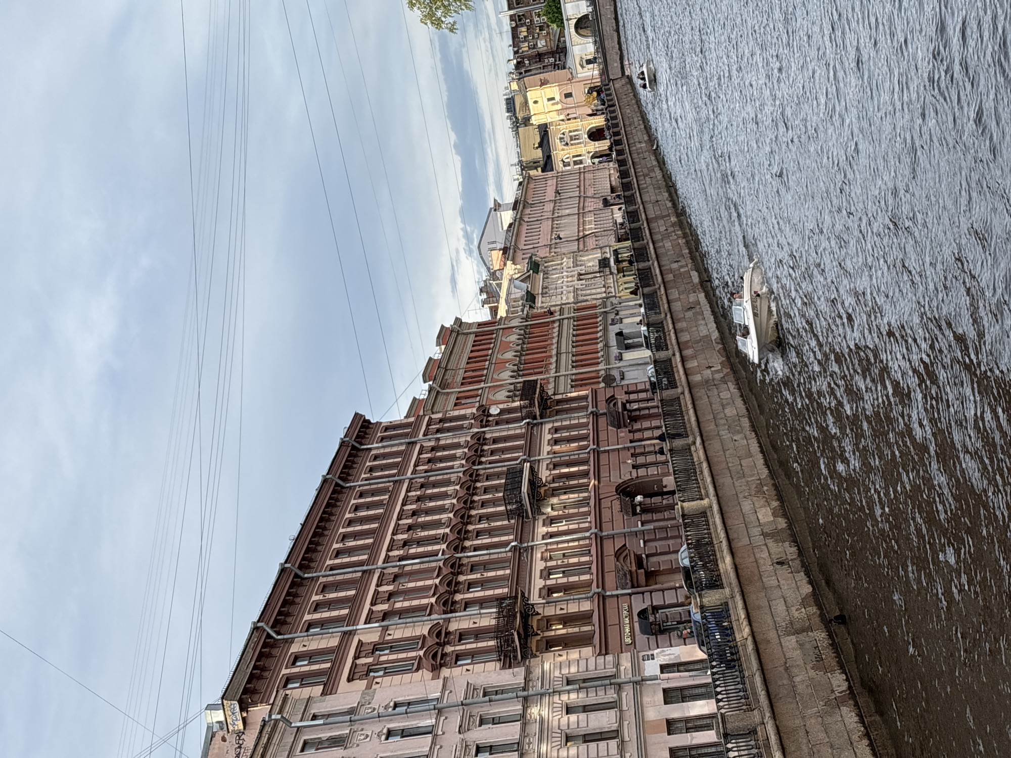 The image, originally oriented sideways, depicts a canal or river scene in Saint Petersburg, Russia, under a cloudy daytime sky. On the left, a long, multi-story building with elaborate architecture dominates the frame. Its facade features reddish-brown and lighter cream-colored sections, numerous rectangular windows, and decorative elements like ornate sills and small balconies. A complex network of external utility pipes runs along the building's exterior. Near the bottom, a sign on the building reads "ЦВЕТОЧНАЯ МАСТЕРСКАЯ" (Tsvetochnaya Masterskaya), which translates to "Flower Workshop" or "Flower Shop." Further down the embankment, other buildings with varied architectural styles are visible.
The upper portion of the sky is light grey with visible clouds, and several thin overhead wires stretch across the frame, converging towards the left. On the right, a wide waterway is filled with rippled, dark grey water. A sturdy stone embankment, topped with a dark metal railing, separates the buildings from the canal. Several small, open boats are visible on the water, some positioned close to the embankment, with small figures of people discernible within them, suggesting leisure activity or transport. The scene appears calm, under the diffused light of what seems to be a cloudy day.