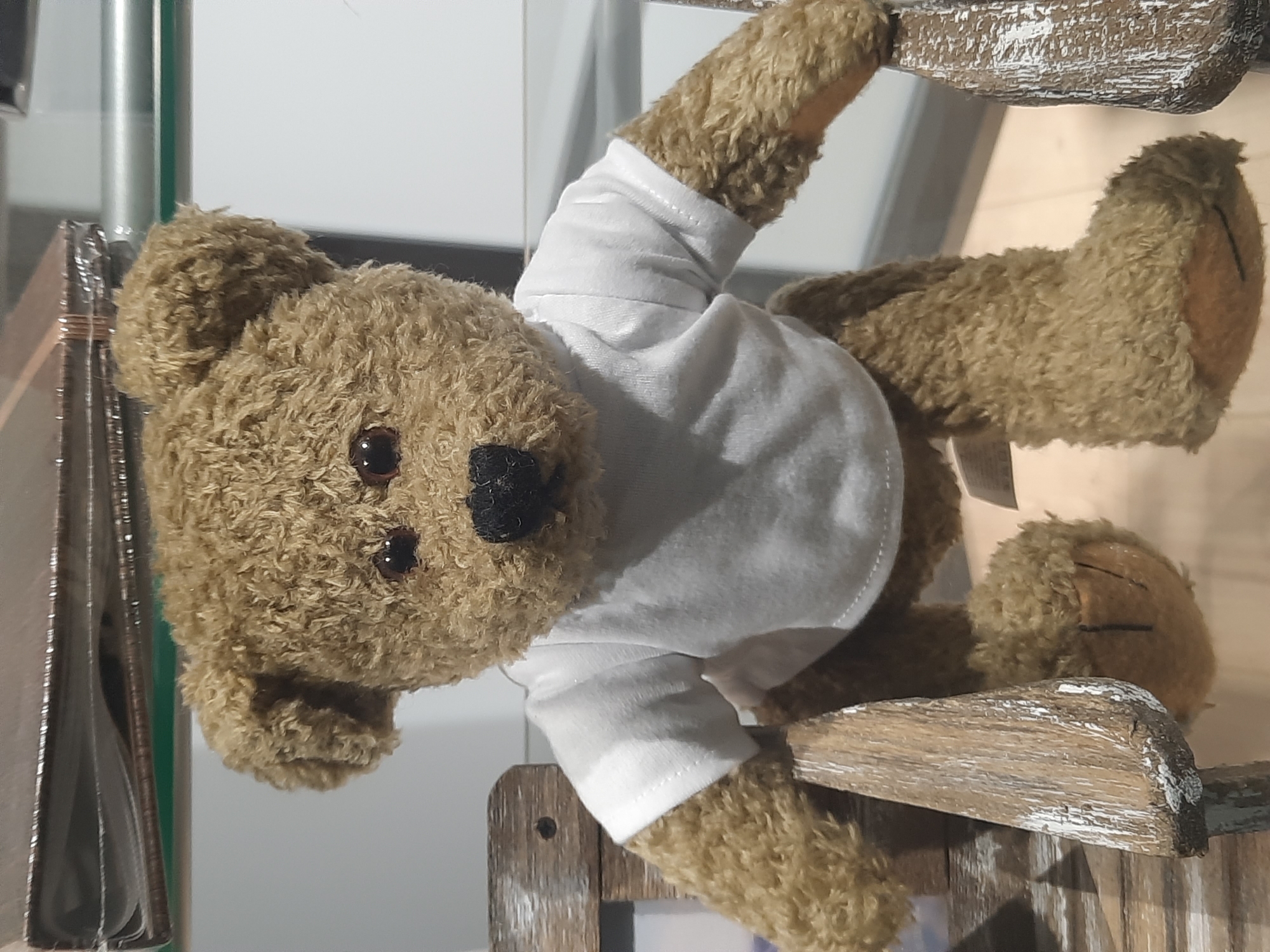 A plush teddy bear, facing forward and seated on a wooden bench, is depicted in a frontal shot. The bear is wearing a plain white t-shirt. Its fur is a light brown, and it has dark, round eyes and a black nose and mouth. The bench appears to be aged, with visible wood grain and some white paint chipping. The background consists of shelves with what appear to be books or photo albums and a plain white wall. The overall lighting is neutral. This image was taken in the city of Rosenheim, Germany.