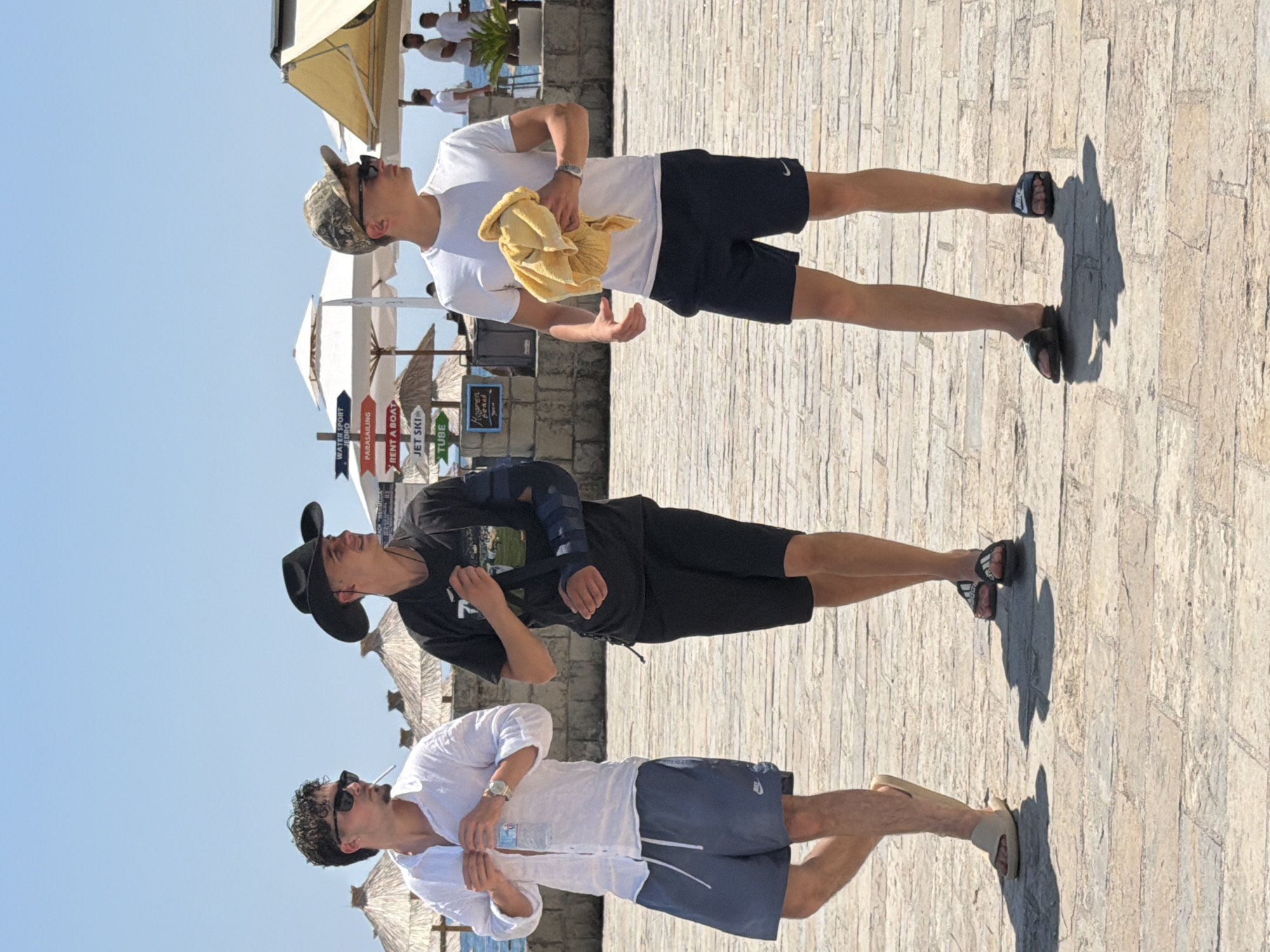 Three young men are walking on a stone-paved promenade in Budva, Montenegro, with the beach and sea in the background under a clear blue sky. The man on the left wears a white unbuttoned linen shirt over a dark undershirt, blue shorts, and light-colored slides. He holds a clear plastic water bottle. The middle man is dressed in a black t-shirt and black shorts, wearing a black cowboy hat and a dark blue arm sling on his right arm. The man on the right is in a white t-shirt, dark shorts, and a camouflage baseball cap. He wears sunglasses and holds a yellow towel. Beach umbrellas and signs indicating water sports are visible behind them, along with other beachgoers and buildings in the distance.