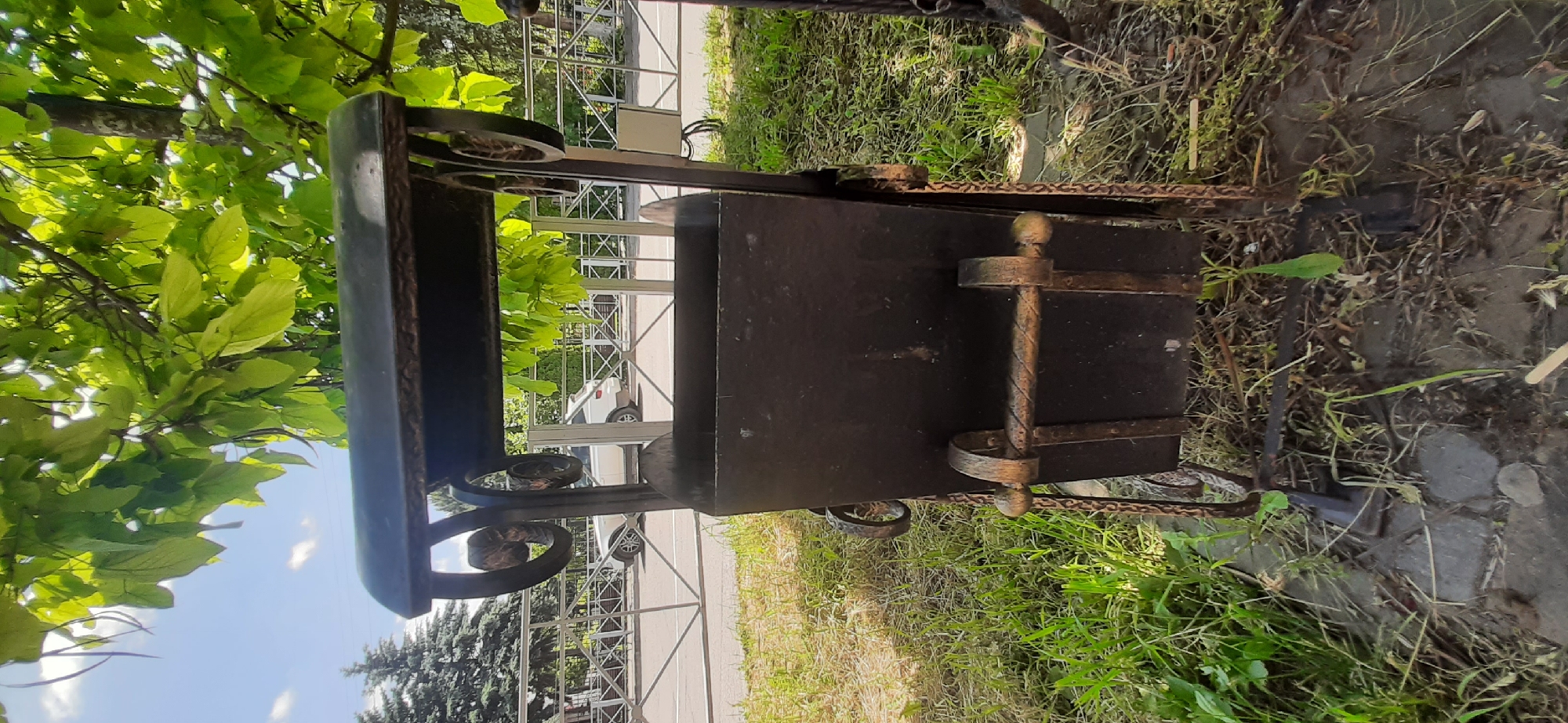 The image shows a black metal trash can with decorative scrollwork on its stand. The trash can is situated outdoors, with green grass and some gravel in the foreground. Behind the trash can, there is a metal fence and a road where a white car is visible. Trees with green leaves are overhead, casting shadows and providing shade. The sky is blue with some white clouds. The location is Yessentukskaya, Russia. There are no people or actions depicted in the image.