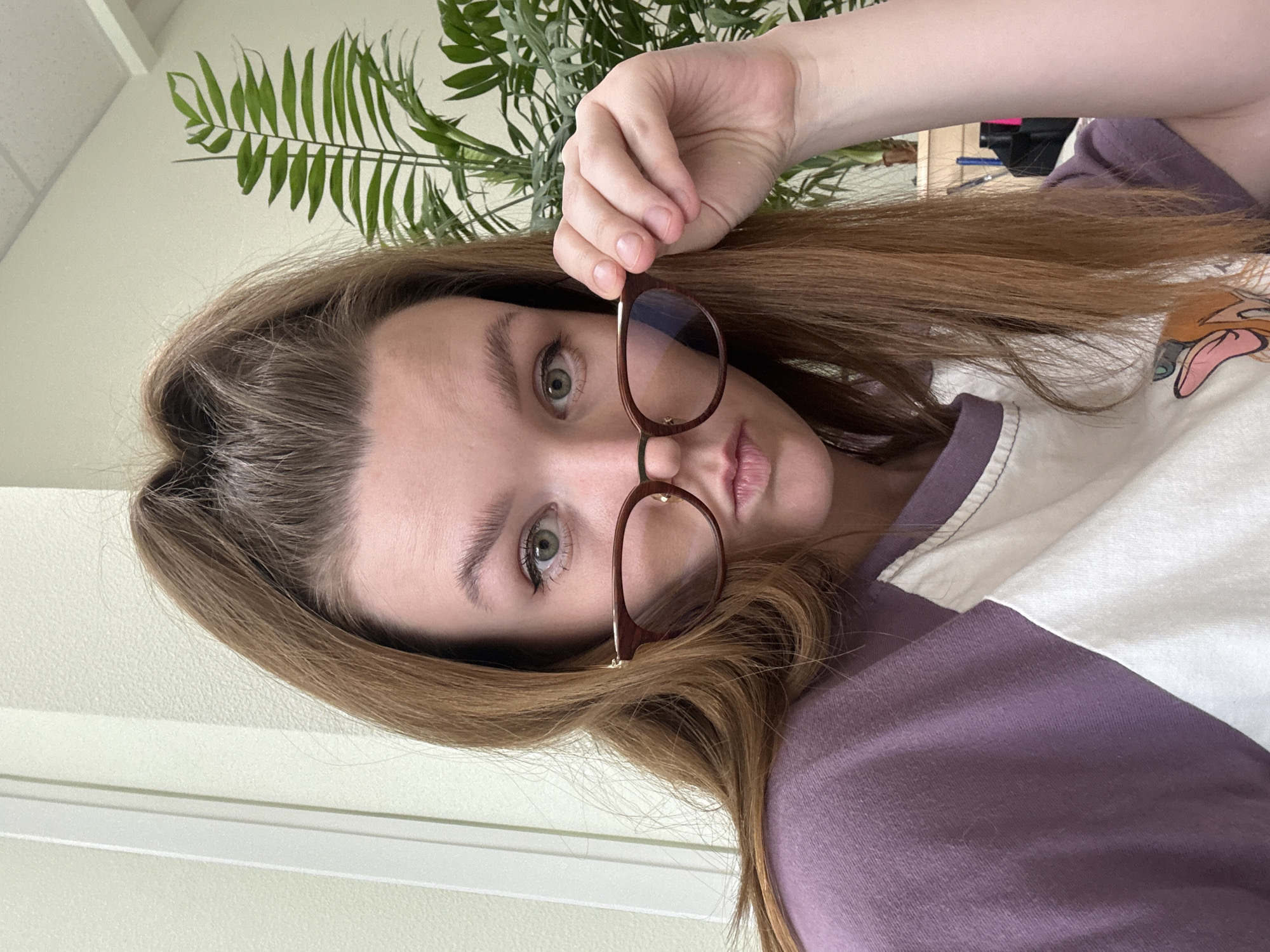 A close-up, eye-level indoor photograph, rotated 90 degrees clockwise, features a young woman looking directly at the camera. She has long, reddish-brown hair and bright green or hazel eyes. Her lips are slightly parted in a soft pout. She is holding a pair of eyeglasses with thick, dark brown or maroon frames just below her eyes, as if peering over them or in the process of taking them off or putting them on. Her right hand is visible, gently gripping the glasses.
She is wearing a two-toned garment, likely a t-shirt or sweatshirt, with the upper right portion being white and the lower left being a muted purple or mauve. A partial illustration of a cartoon deer character, resembling Bambi, is visible on the white section of her top.
The background consists of a plain, light-colored wall (off-white or cream) and a vibrant green leafy plant, possibly a houseplant like a palm or fern, partially visible in the upper left corner. The lighting appears to be ambient indoor light. The overall mood of the image is neutral and direct, with the subject engaging the viewer with her gaze. There is no discernible text. This indoor setting is contextually located in Izhevsk, Russia.