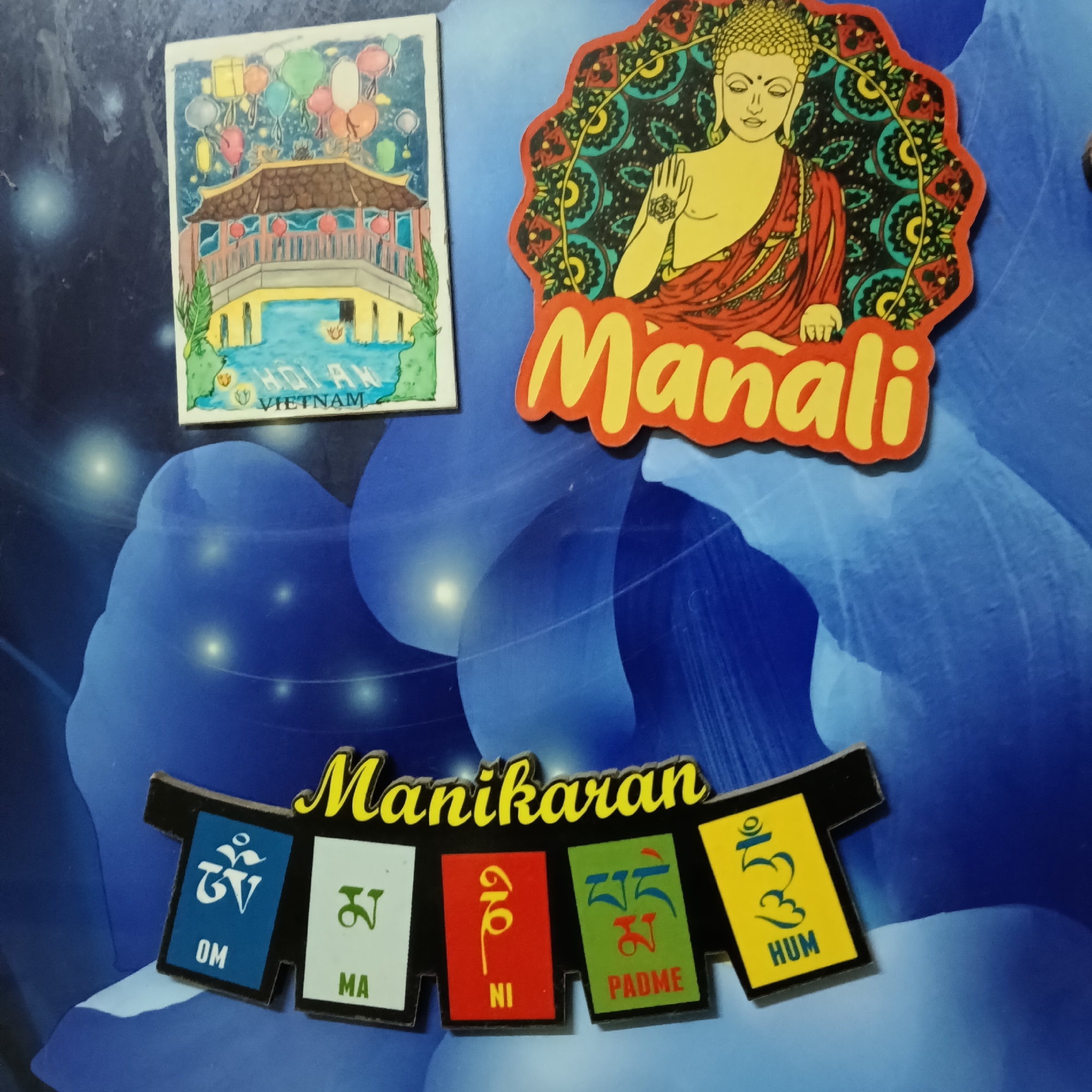 The image shows several refrigerator magnets attached to a blue, textured surface, likely a refrigerator door in a residential kitchen.
In the upper left is a rectangular magnet depicting a bridge and traditional buildings with lanterns, labeled "HOI AN VIETNAM". To the right of this is a circular magnet featuring an image of Buddha in meditation with intricate patterns and the word "Manali" in yellow text with an orange outline.
Below these, a prominent magnet with a black background and a wavy top edge is adorned with Buddhist prayer flags. The text "Manikaran" is written in yellow cursive script across the top. Each flag has a distinct color and a symbol or text:
- The first flag is blue with a white symbol and the text "OM" in white.
- The second flag is white with a green symbol and the text "MA" in black.
- The third flag is red with a yellow symbol and the text "NI" in red.
- The fourth flag is green with a blue symbol and the text "PADME" in white.
- The fifth flag is yellow with a blue symbol and the text "HUM" in black.
The blue surface has a slightly iridescent or textured appearance with subtle light reflections, suggesting it's a common household appliance. The overall scene is a collection of travel-related souvenirs.
There is no visual information in the image to suggest this location is in Bengaluru, India, or any specific city for that matter. The objects are static and there are no people present.