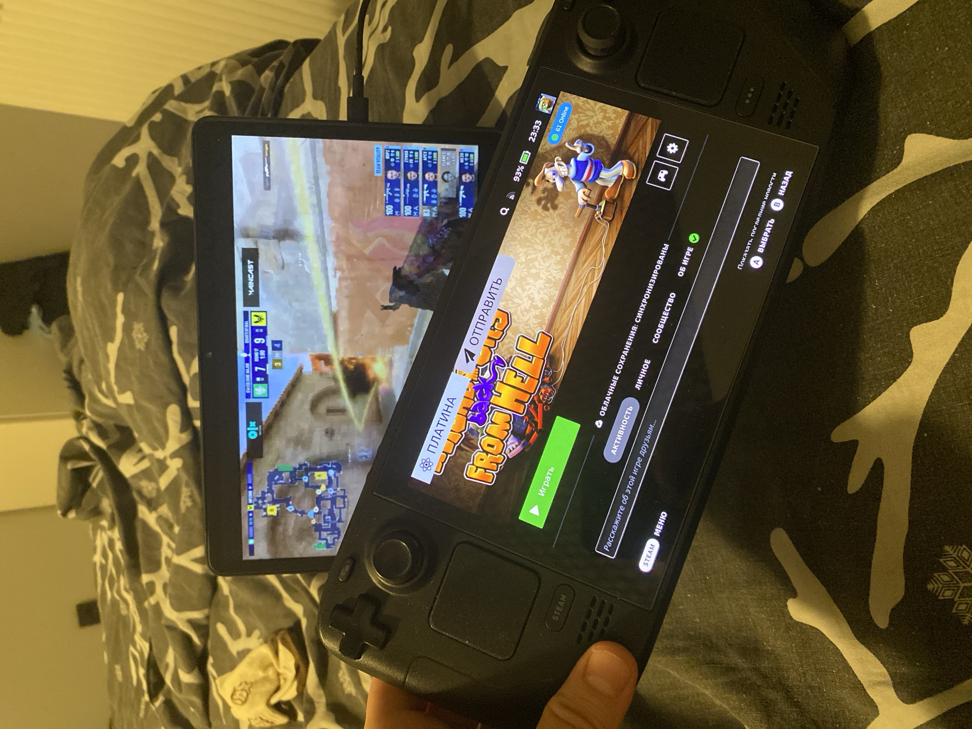 The image shows a person holding a black Steam Deck gaming console in their hands. The Steam Deck is displaying a game's menu, featuring the title "FROM HELL" prominently. The game appears to be a shooter, as indicated by the text "АКТИВНОСТЬ" (Activity) and "ОБЛАЧНЫЕ СОХРАНЕНИЯ: СИНХРОНИЗИРОВАНЫ" (Cloud Saves: Synchronized). In the background, a tablet is propped up, showing a live esports match, likely a first-person shooter given the on-screen graphics and player statistics. The overall environment suggests a casual setting, possibly a bedroom, with bedding visible in the background. There is no indication of the city of Łódź, Poland in the image.