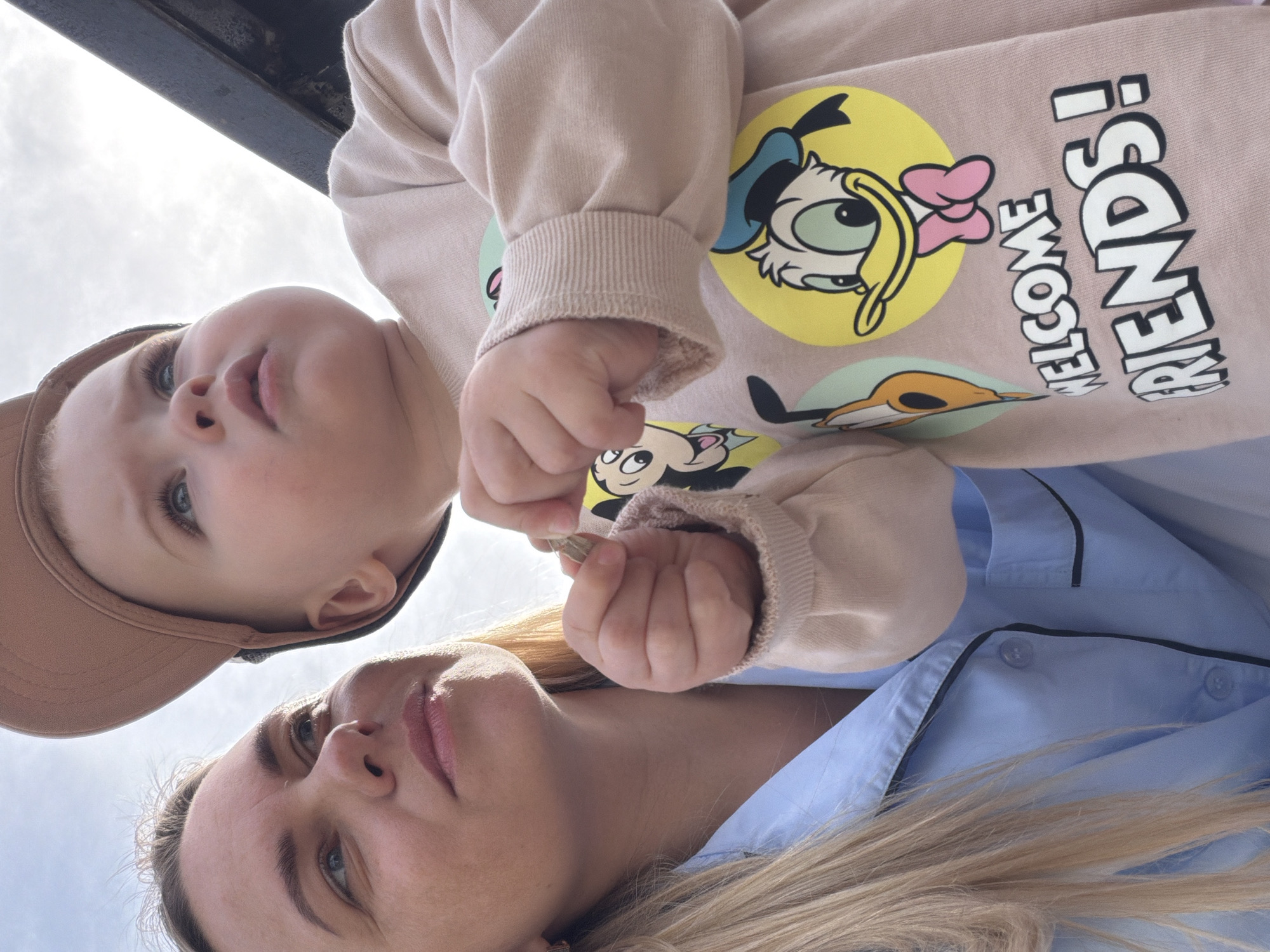 The image features two individuals, a young child and an adult woman, captured from a low-angle perspective looking upwards against a bright sky.
The child, positioned higher in the frame, wears a light beige baseball cap and a pale peach long-sleeved sweatshirt. The sweatshirt is adorned with cartoon prints, notably a yellow circular graphic featuring Donald Duck's head and the text "WELCOME FRIENDS!" written vertically in white letters with a dark outline. Another smaller cartoon character, possibly Mickey Mouse, is visible lower on the shirt. The child has light-colored eyes and a focused or curious expression, with their right hand partially visible, holding a small, metallic, key-like object.
Below the child is an adult woman, visible from her forehead and eyes downwards. She has blonde hair and light-colored eyes, with a neutral to slightly serene expression. She wears a light blue collared shirt with dark piping along the collar and front placket. Her hair flows over her shoulder and parts of her shirt.
The background primarily consists of a bright, overcast sky, indicating daytime. A dark, linear structure, possibly a beam or railing, is visible along the top edge of the frame. The image captures a quiet, shared moment, likely outdoors, in Votkinsk, Russia.